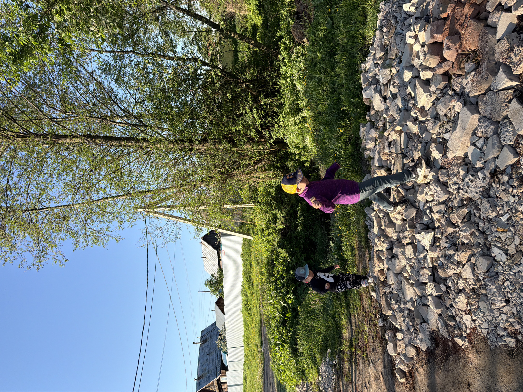 The image depicts an outdoor scene, likely taken in a rural area, with two children present. The location is reported to be in the city of Chaikovsky, Russia, but no visual elements in the image confirm this.
The setting includes a dirt road, grass, trees, and some buildings in the background. A pile of loose rock or rubble lies beside the road. There are power lines running above the buildings.
The two children are positioned near the pile of rocks. One child is wearing a purple jacket, jeans, and a yellow and blue cap. The other child wears a black top with a white pattern and a blue cap. The children appear to be standing and interacting with the rocks. The posture of the child with the yellow and blue cap gives an impression of eating something.
The image is oriented sideways. The sky is visible in the lower left portion. The lighting suggests it is daytime.