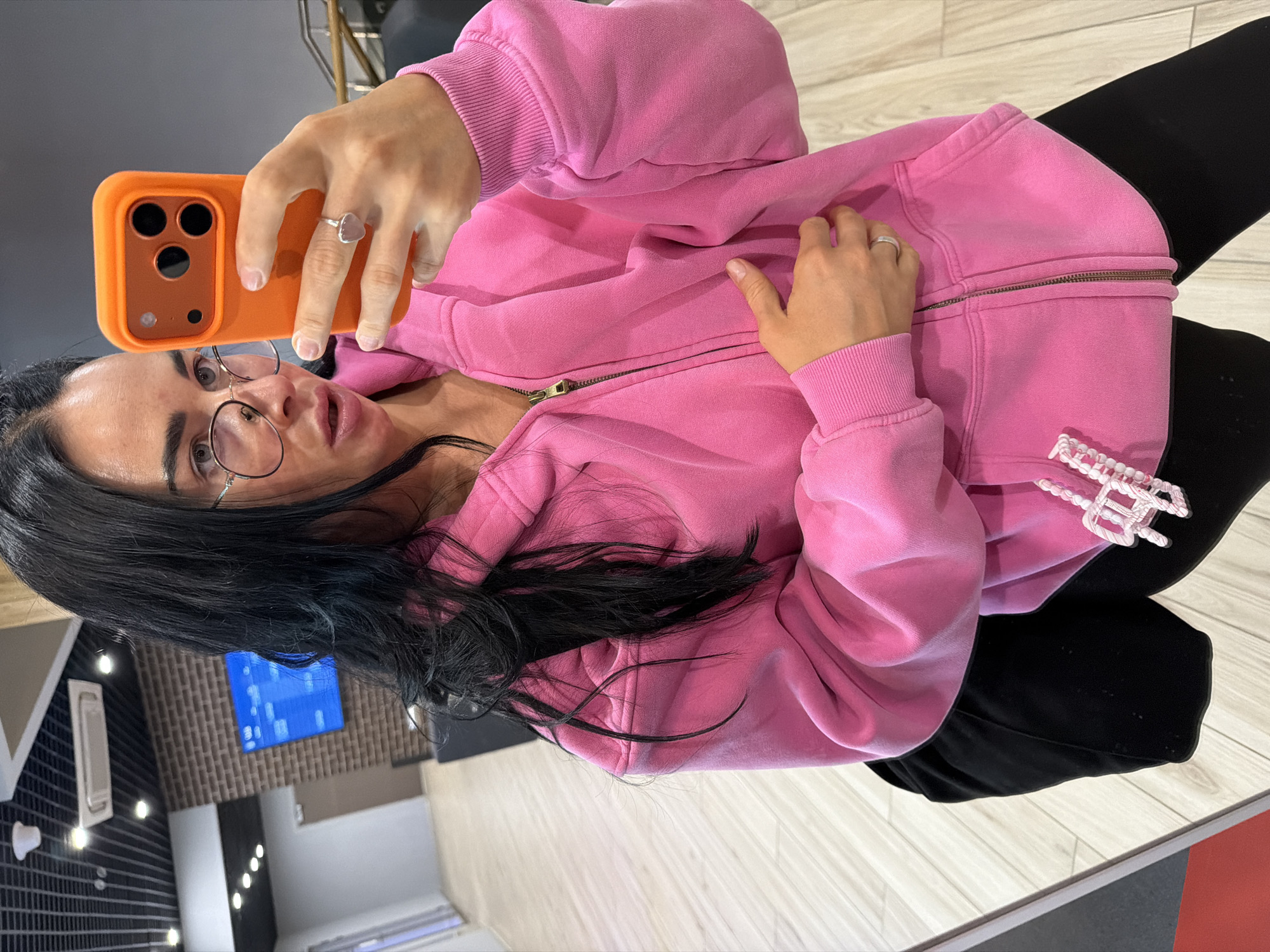 The image displays a woman taking a selfie in a mirror or highly reflective surface, captured from a tilted perspective. She has long, dark hair and wears round, thin-framed glasses. Her mouth is slightly open, and she appears focused on the camera.
She is dressed in a bright, fuchsia pink zip-up hoodie with ribbed cuffs and a metal zipper, and black leggings or pants. On her right hand, which holds an orange-cased smartphone featuring multiple cameras, she wears a silver-colored ring with a light-colored, possibly pink or translucent, heart-shaped stone. Another simple silver-colored ring is visible on her left hand, which rests near her chest on her hoodie. Attached to the lower right side of her hoodie or pants is a decorative white and pink beaded or braided accessory.
The background, seen in the reflection, indicates an indoor setting. It features light-colored flooring that resembles wood planks, and parts of a wall with a textured, possibly tiled, or patterned design. A blue screen or monitor displaying white elements is visible in the background, along with several ceiling lights. The overall environment suggests a modern commercial space, such as a gym, store, or salon, rather than a home. The lighting is artificial, and no specific time of day or external location cues for Saint Petersburg, Russia, are discernible from the image itself. No clear text is visible.