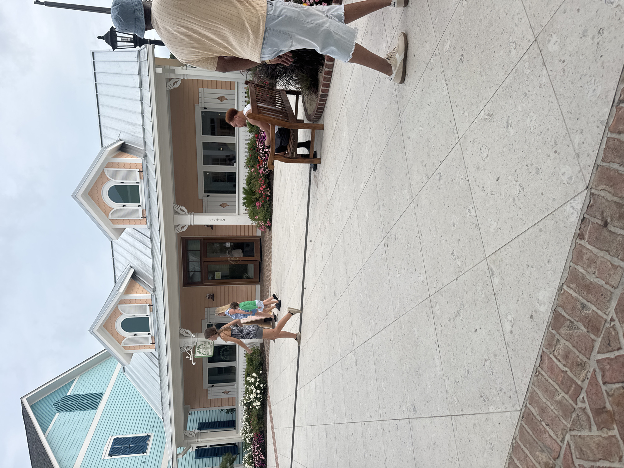 The image depicts an outdoor scene in Myrtle Beach, United States of America, likely at a retail or entertainment complex, as suggested by the architecture of the buildings. In the foreground, a person wearing a light tan t-shirt and light blue denim shorts walks away from the camera, their face not visible. Behind them, a person with red hair sits on a wooden bench, looking to the left. To the left of the bench, several people are walking, including a young woman in a patterned dress and grey shorts, another woman in a blue and white floral dress, and a child in a green shirt. The buildings in the background are brightly colored, with a blue building to the left and a pinkish-orange building in the center. Both buildings have prominent dormer windows and metal roofs. The ground is paved with large square tiles, with a border of brickwork in the lower left corner. There are planters with flowers in front of the pinkish-orange building. A street lamp stands to the right of the pinkish-orange building. The sky is overcast.