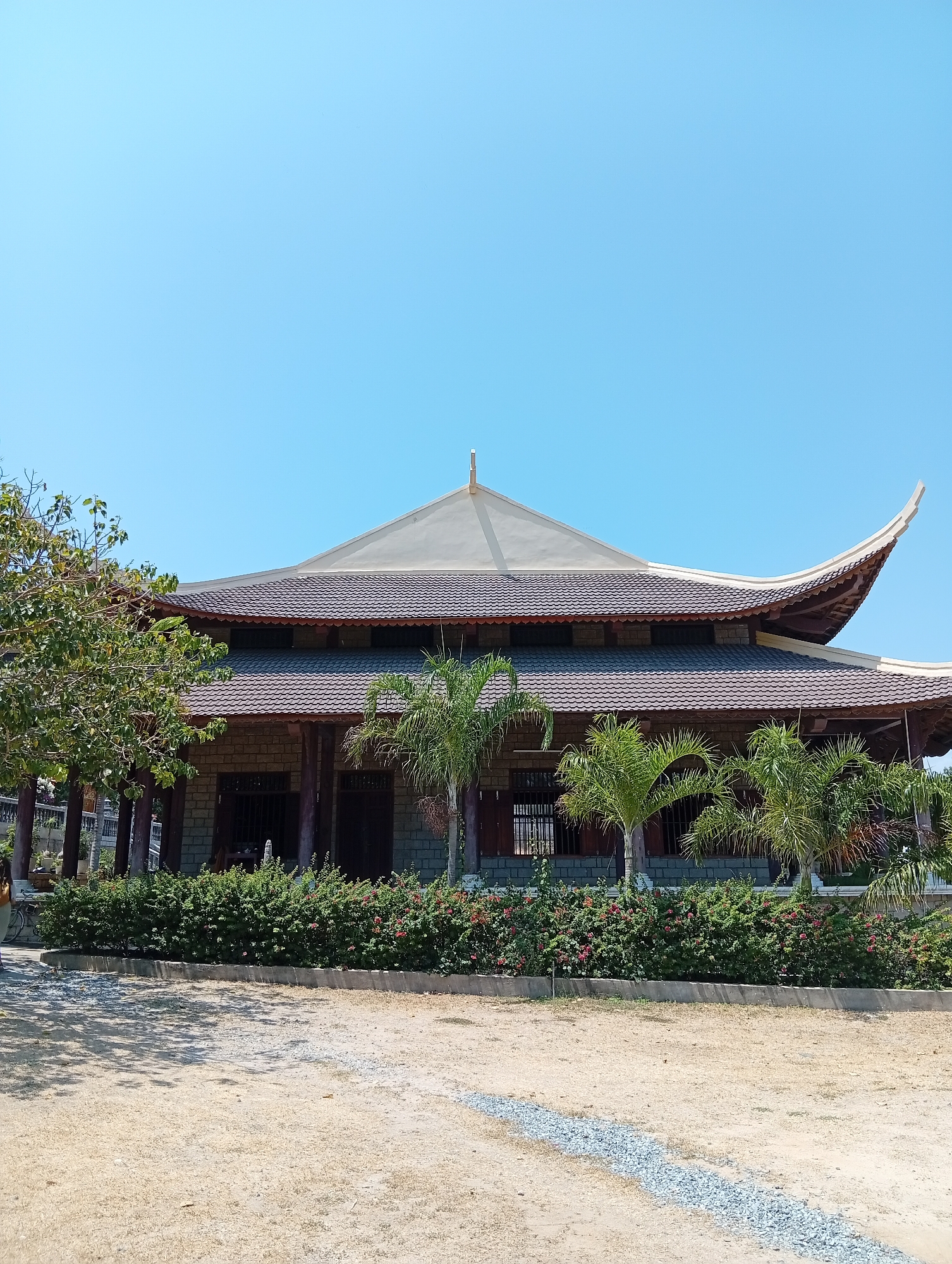 The image shows a traditional Vietnamese building in Khánh Hải, Vietnam. The building has a multi-tiered, curved roof with brown tiles and a light gray peak. The structure features stone walls and wooden pillars, with windows partially visible behind the pillars. Lush green bushes with red flowers line the front of the building, interspersed with small palm trees. A large tree with green leaves is visible to the left. The foreground is a sandy area with a strip of gravel leading towards the building. The sky is clear and blue.