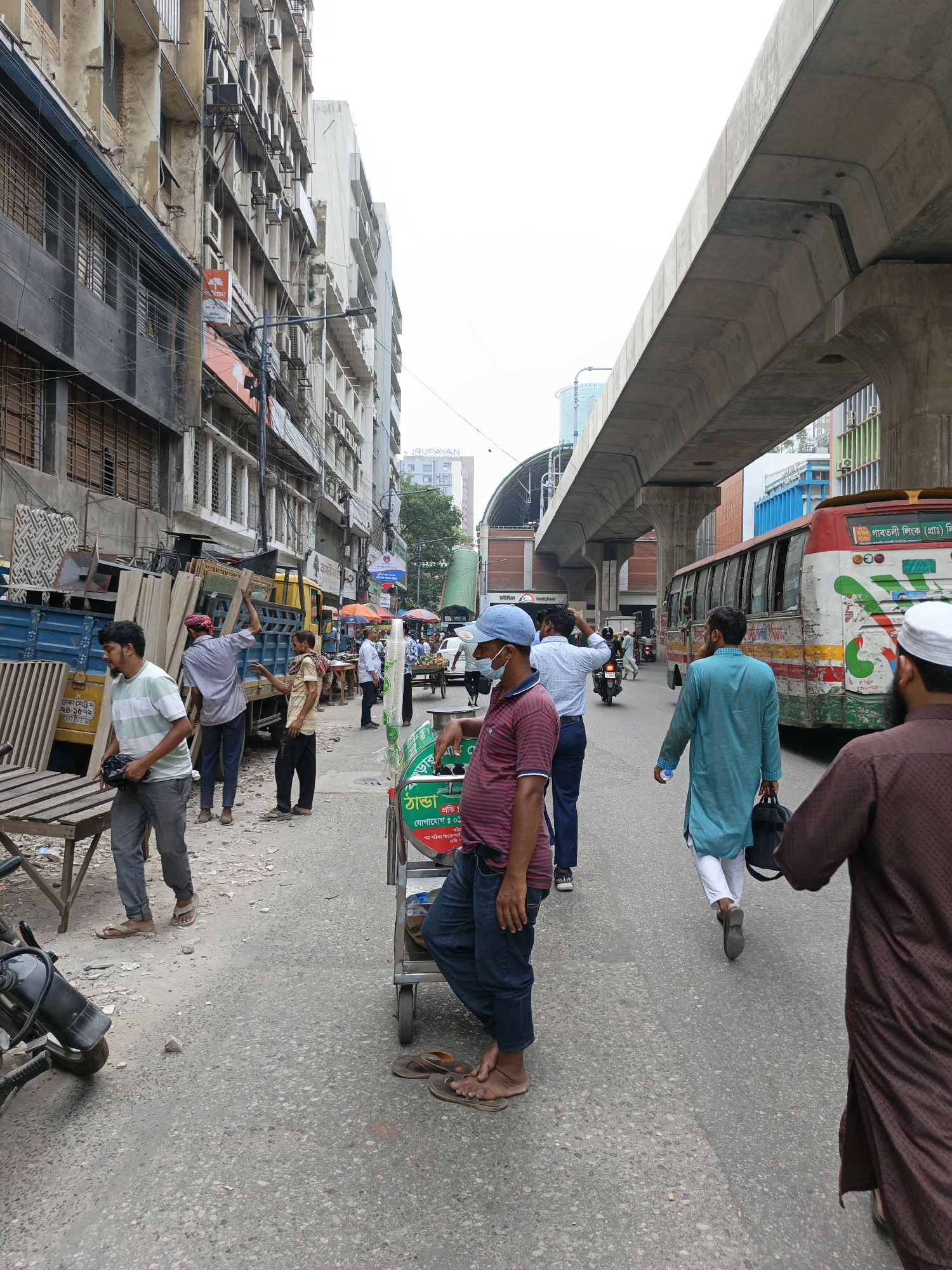 The image depicts a street scene in Dhaka, Bangladesh. The street is congested with pedestrians, vehicles, and vendors. The buildings on the left are old and weathered, with many air conditioning units installed. On the right, an elevated concrete railway structure dominates the view. A brightly colored bus is visible. Several men are walking on the street, one wearing a blue kurta and carrying a black bag. Another man in the foreground is wearing a striped shirt, jeans, and a blue cap, leaning against a green cart. The ground is asphalt, showing signs of wear and tear. Some men are unloading wooden planks from a blue truck. The overall impression is of a busy, somewhat chaotic urban environment. "Motijheel" is labeled on a nearby building.