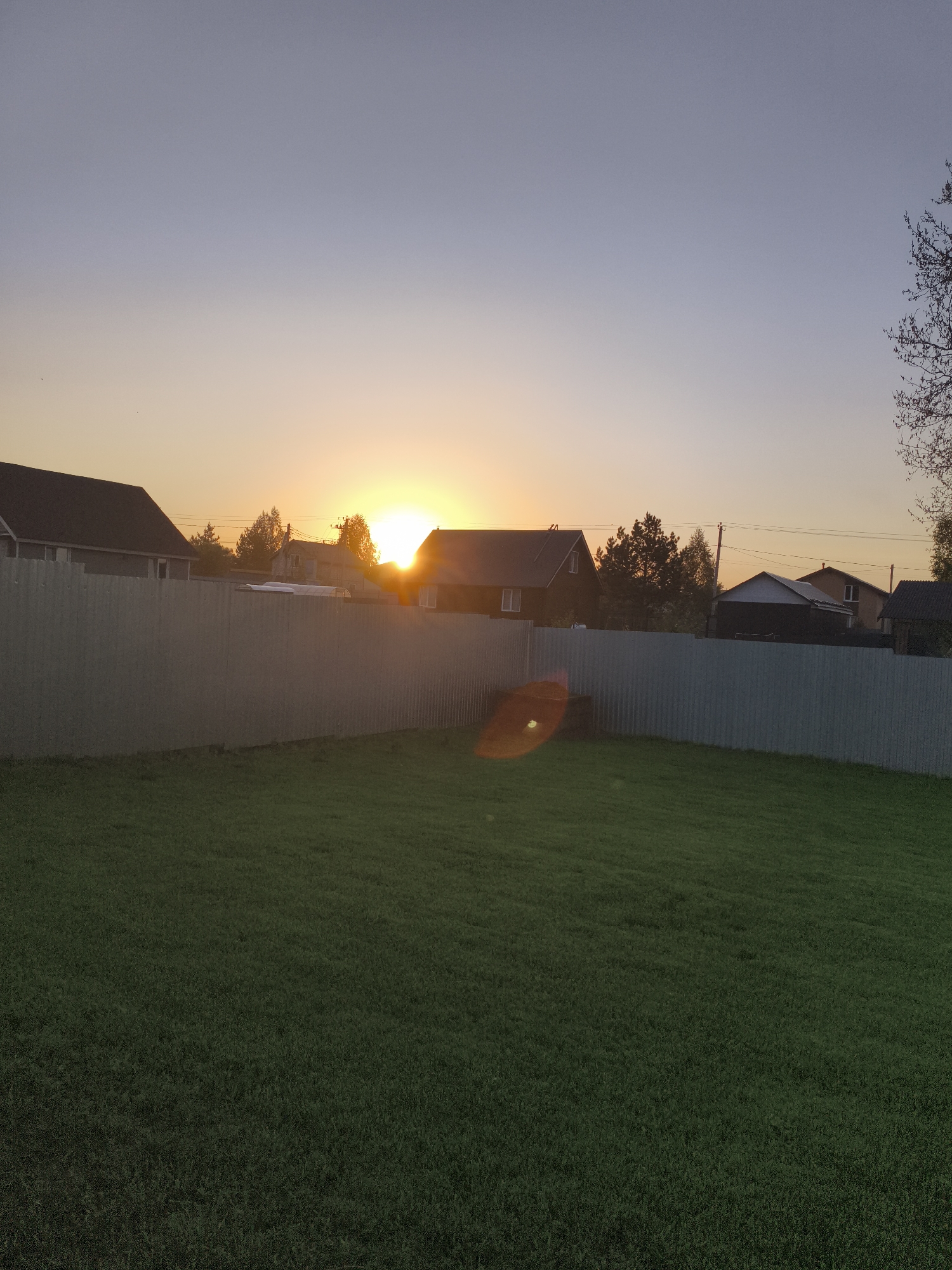 The image depicts a fenced backyard in the Russian settlement of Русский Вожой. The setting sun is visible above the houses and trees, casting a warm light on the scene. A tall, corrugated metal fence encloses the yard, which is covered in green grass. A lens flare from the sun is visible on the fence and the lawn. The neighborhood consists of small houses with various roof styles. Utility poles and wires are also visible against the sky.