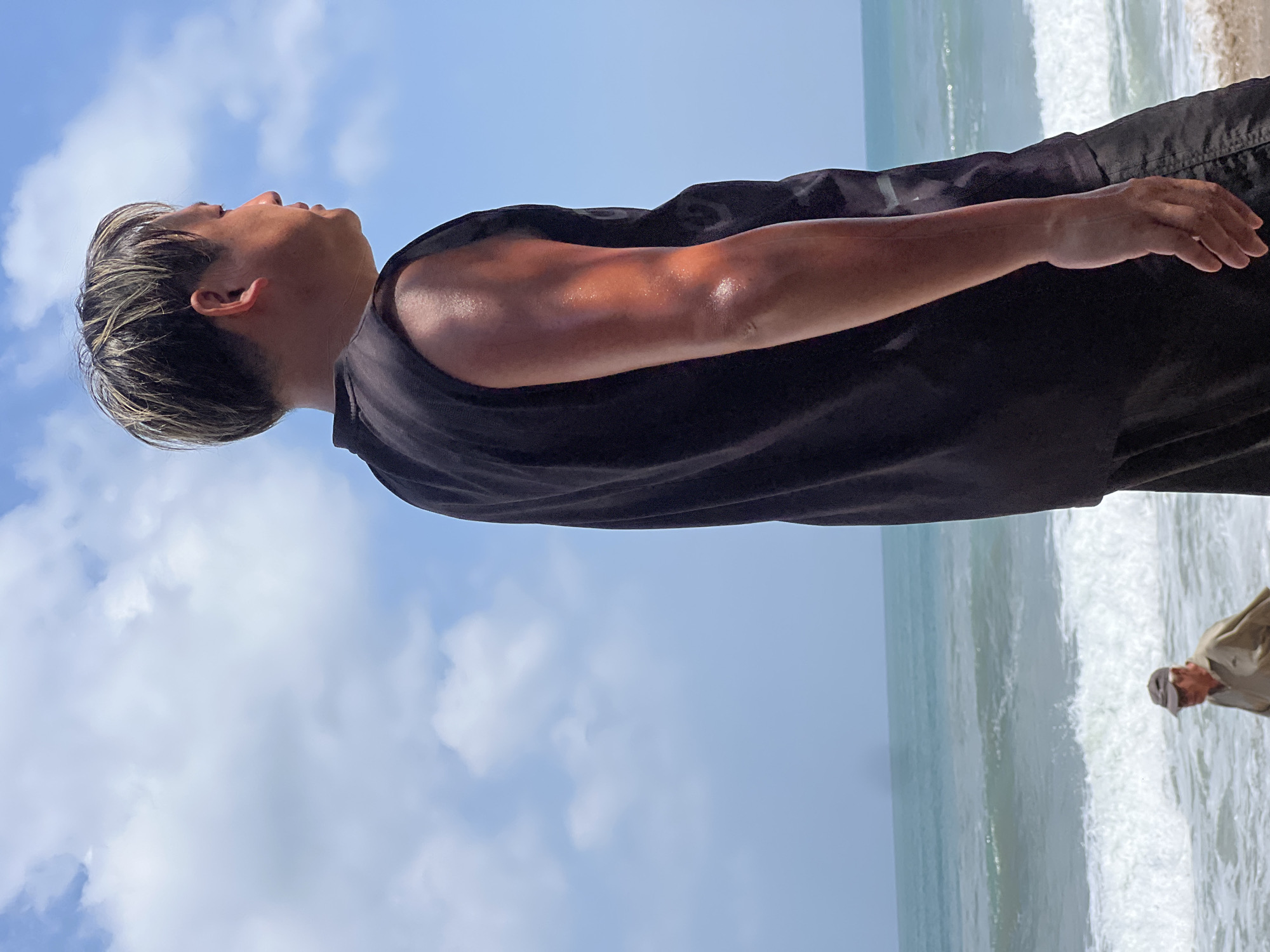A man is seen from the side, facing right, wearing a black sleeveless shirt and black shorts. He has styled hair with blonde highlights. His arm glistens with sweat. In the background, the ocean meets the sky under a partly cloudy blue sky. A smaller figure of an older person wearing a cap and a light-colored shirt walks along the shore, near the breaking waves. The scene appears to be a beach, possibly in Kuta, Indonesia, given the characteristic waves and shoreline.