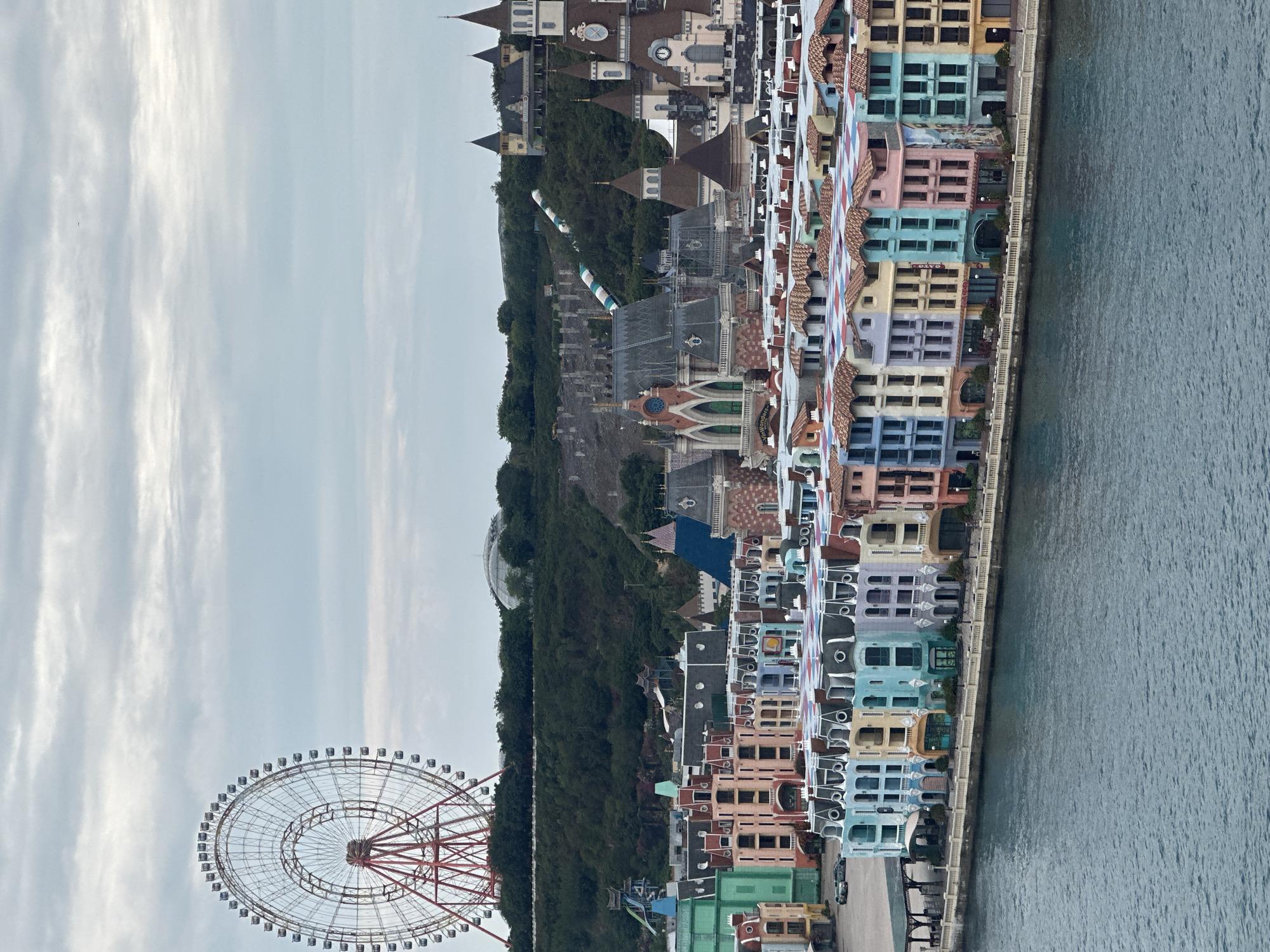 The image presents an overview of a coastal landscape featuring a blend of architectural and natural elements, situated in Nha Trang Ward, Vietnam. A large Ferris wheel with a red support structure occupies the lower left of the frame. The wheel is prominent against the sky, which is filled with wispy clouds.
The center of the image is dominated by a green, densely wooded hill that descends towards the sea. The hill supports a variety of buildings, ranging from castle-like structures with pointed roofs to more modern constructions. A water slide is visible on the hill.
Adjacent to the wooded area, a row of brightly colored, multi-story buildings lines the coast. These buildings feature a variety of colors, including pastel shades of blue, pink, yellow, and green, and have tiled roofs.
The water extends to the right of the frame and appears calm, with subtle variations in color suggesting varying depths. Along the shoreline, there is a visible barrier or wall separating the buildings from the water.