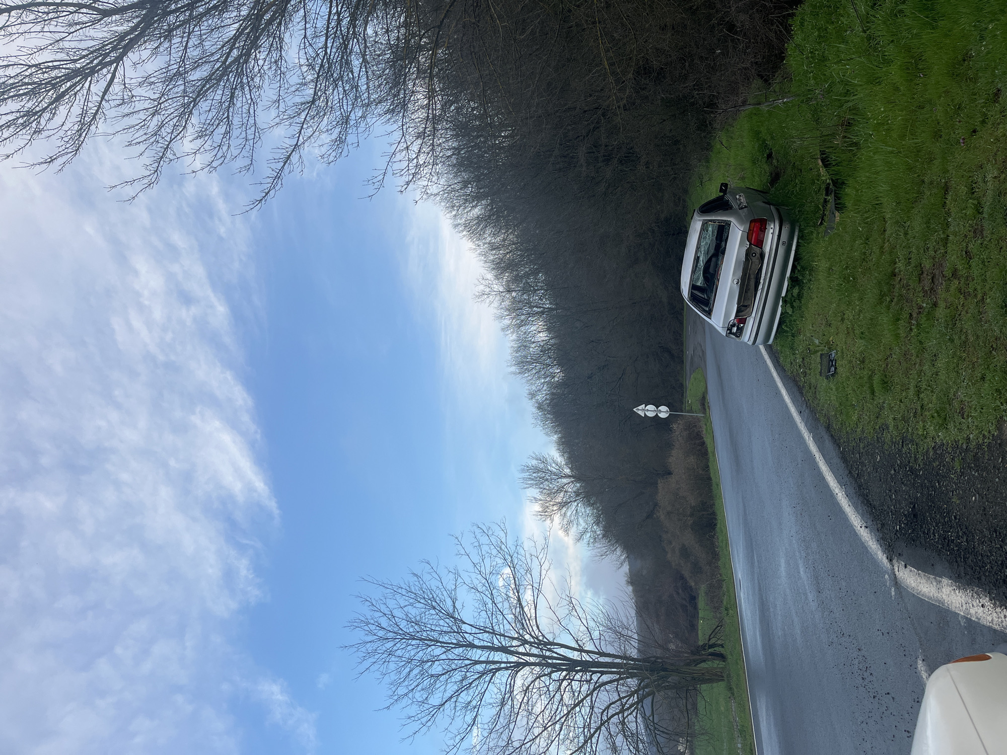 The image shows a silver car crashed off the side of a road in a rural area. The car is positioned on a grassy embankment, partially obscured by trees. The rear window of the car is shattered. The road is paved with a white line marking the edge. The sky is blue with scattered clouds. Trees line both sides of the road. A road sign is visible further down the road. The location appears to be Gabar, Bulgaria.
