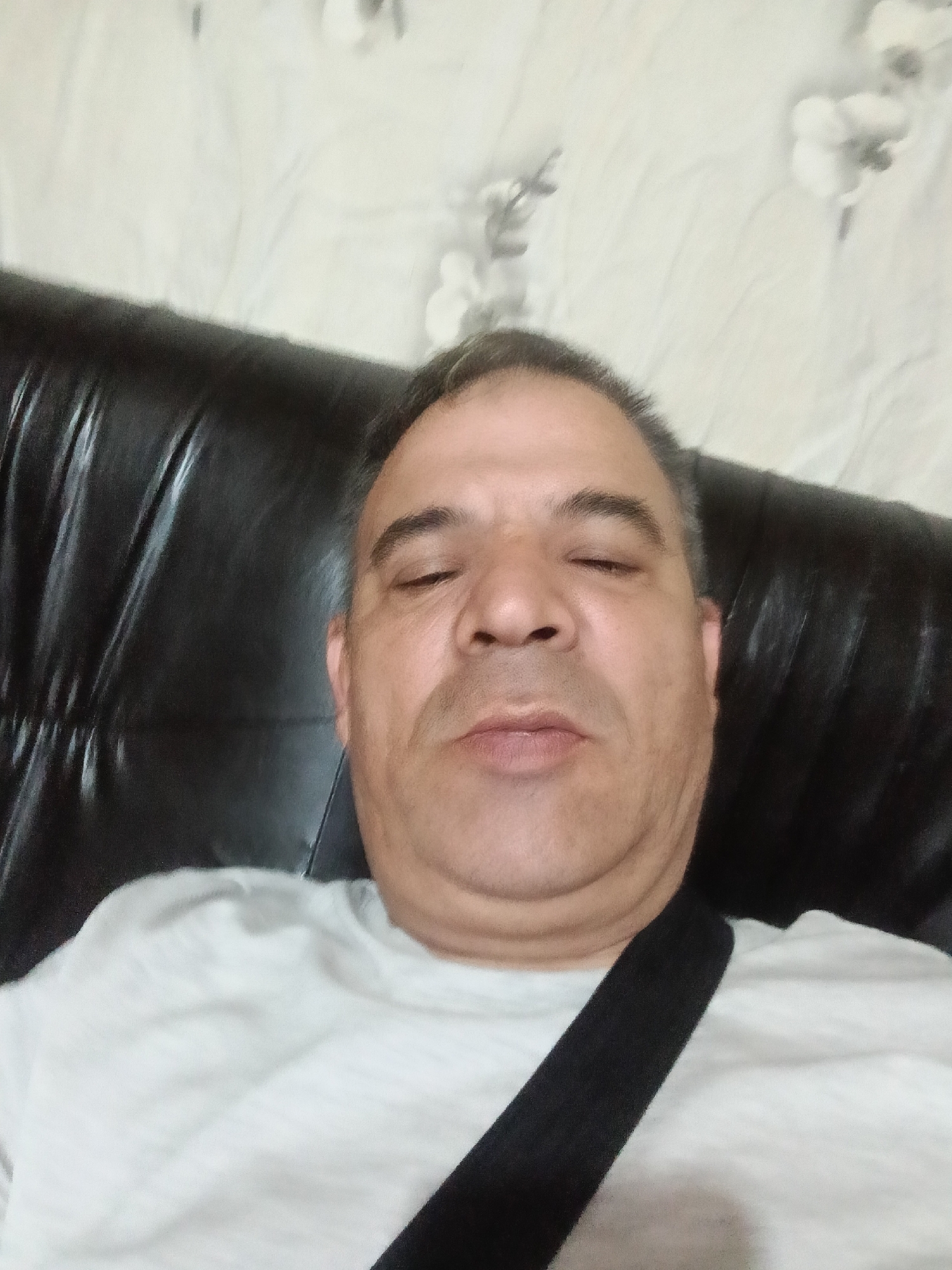 A middle-aged man is captured in a close-up, chest-up portrait, appearing to be in a state of rest or sleep. His eyes are closed, and his expression is relaxed, indicating a moment of repose. He has dark, graying hair and dark eyebrows. He is wearing a light-colored, possibly white, t-shirt, with a black strap visible across his right shoulder and chest, which could be from a bag, a seatbelt, or a brace.
The man is leaning against a dark, possibly black, upholstered surface, which appears to be the backrest of a couch or armchair, characterized by horizontal lines and a plush texture. The background consists of a light-colored wall adorned with subtle, patterned wallpaper, featuring faint floral or abstract motifs in shades of white and light gray. The lighting suggests an indoor environment. The overall scene conveys a sense of calm and relaxation, likely within a home setting in Vetovo, Bulgaria. No text is visible in the image.