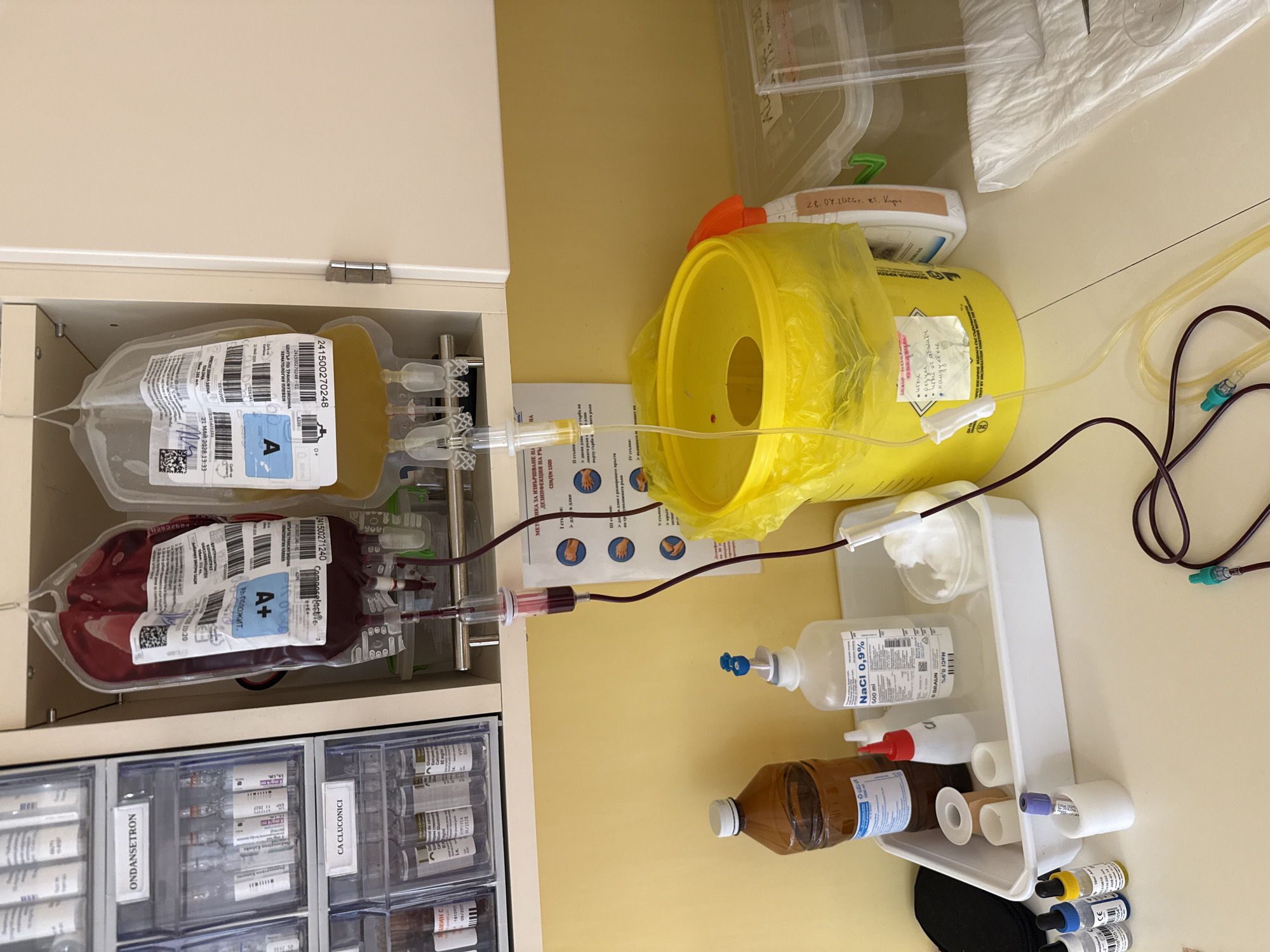 The image shows a medical setting, likely a room in a hospital or clinic in Pleven, Bulgaria. A cabinet contains two blood bags, one with dark red blood labeled "A+" and another with yellow plasma labeled "A," both from the Center for Transfusion Hematology Pleven. Tubes connect the blood bags to a transfusion setup, including a saline solution (NaCl 0.9%) and a sharps container, used for disposing of needles. The cabinet drawers are labeled "Ondansetron" and "Ca Cluconici," referring to medications. The photograph captures a sterile environment with medical supplies readily available for blood transfusion or related procedures. There's a date, "28.07.2026 г. Кт. Кирил" written on a container. A chart depicting hand sanitizing steps is visible. Several vials of medication are also visible in the cabinet drawers.