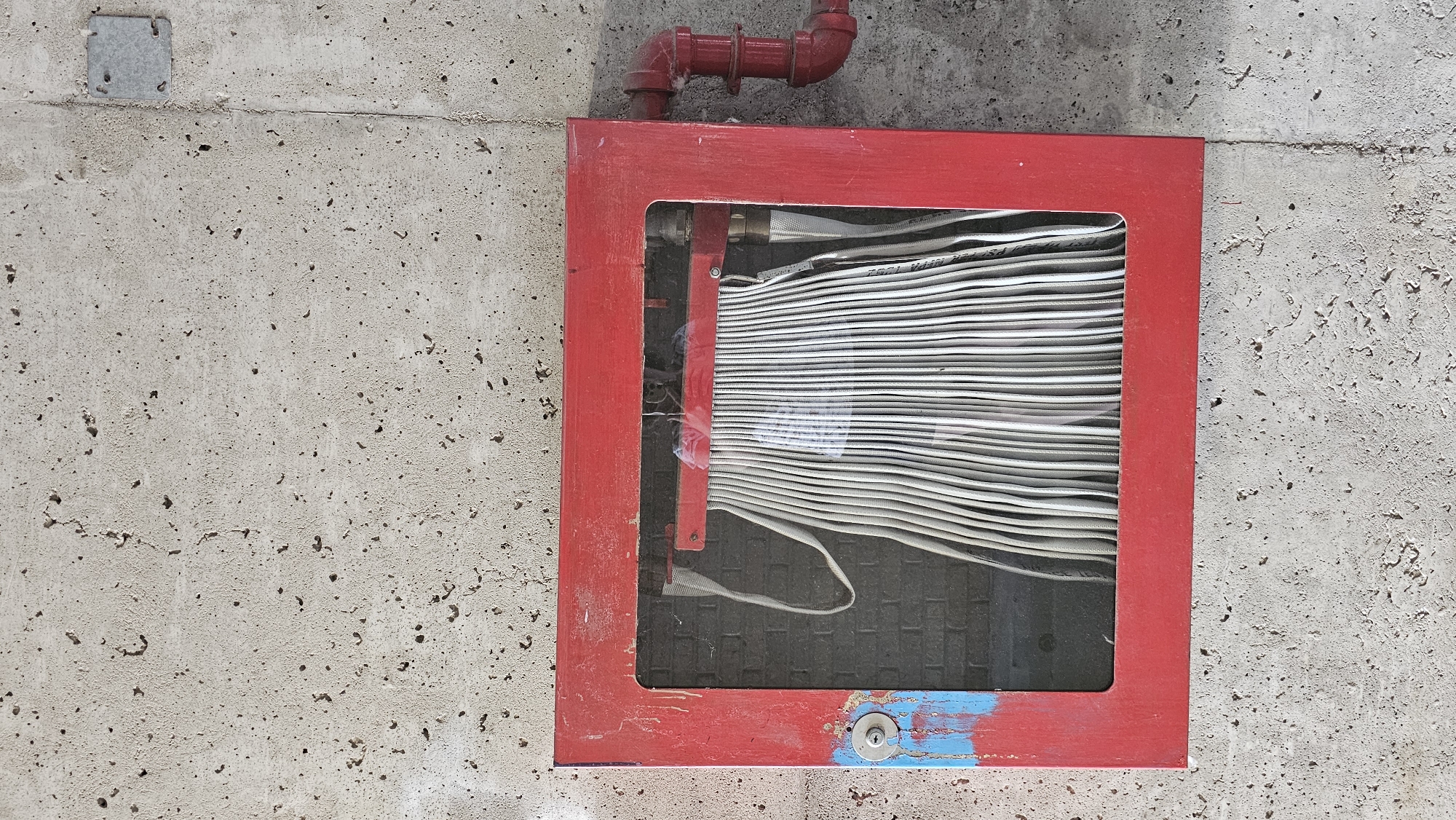 A red metal fire hose cabinet is mounted on a concrete wall. The cabinet has a clear glass front, revealing a white fire hose coiled inside. The hose is neatly folded, with its loops creating a uniform pattern. To the right of the cabinet, a red pipe with elbow joints connects to the wall. The concrete wall has a rough, textured surface with visible imperfections and small dark pits. A metal plate is affixed to the wall to the upper right of the cabinet. The lower left corner of the cabinet shows signs of wear, with blue paint over a previous layer of red paint. The scene appears to be outdoors, given the texture and appearance of the concrete, and likely located in Oakville, Canada. There are no people present in the image.