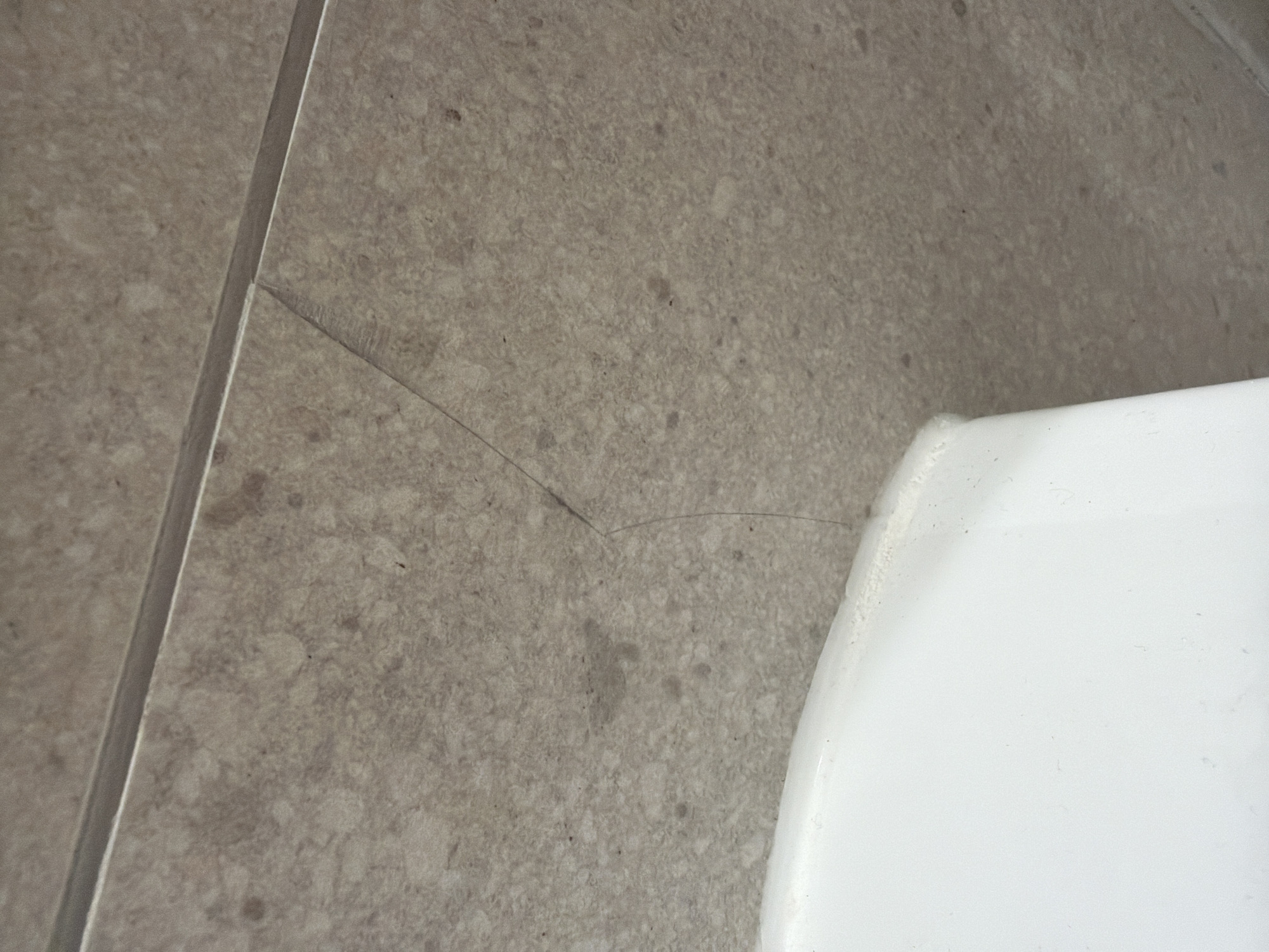 The image shows a close-up of a cracked tile, likely in a bathroom or kitchen setting, with a portion of a white porcelain object, possibly a toilet tank, visible in the lower left corner. The tile is a light brown speckled pattern, with a dark grout line running horizontally across the top. A thin, hairline crack extends diagonally from the grout line downwards into the tile. The crack appears to be relatively fresh, with no visible debris or discoloration within it. The lighting suggests an indoor environment, possibly with natural light coming from above. The scene is devoid of any human presence or activity. The location is not specified.