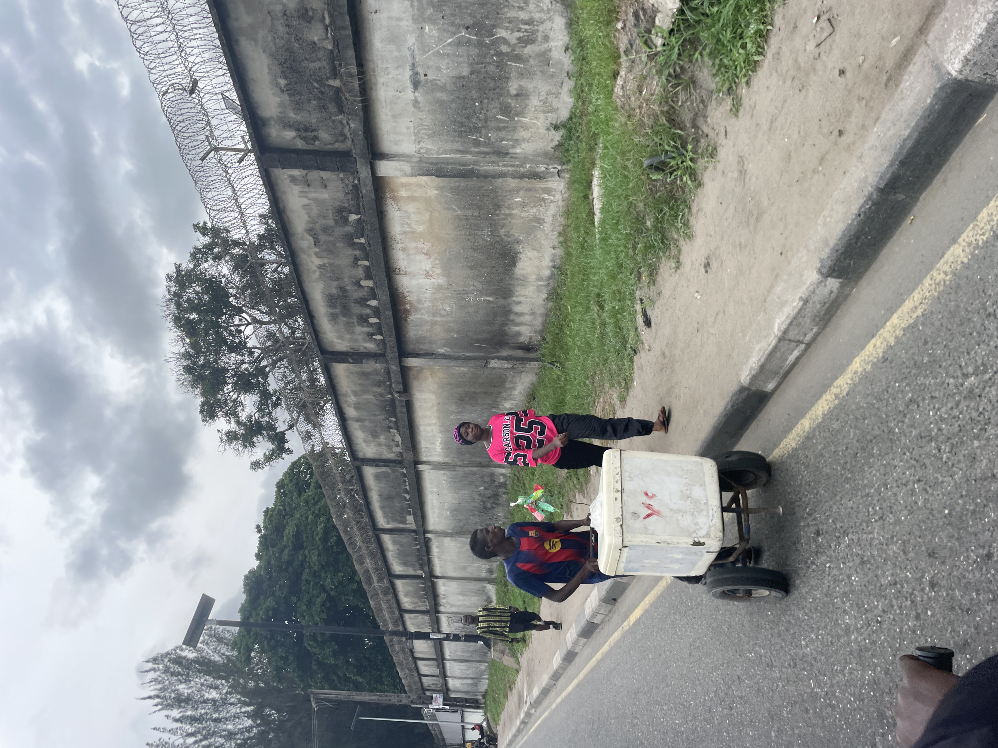 The image depicts an urban scene likely in Lagos, Nigeria, characterized by a high concrete wall topped with barbed wire. Two individuals are present: one pushing a small, wheeled cart with a white container on top, and another standing on the sidewalk in a pink shirt. A third individual is walking on the sidewalk in the background. The environment consists of a grey sky, a concrete wall with barbed wire, a sidewalk with grass, and a paved road. The cart is in motion on the road.
