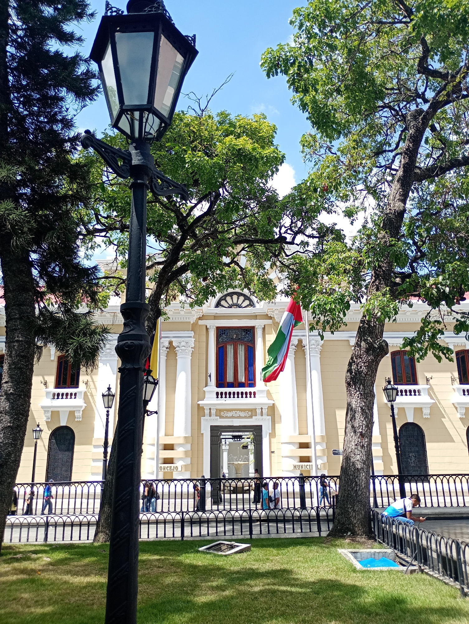 The image captures a street scene in Caracas, Venezuela, centered on the Municipal Council building. The building is a yellow and white structure with classical architectural elements, including columns, balustrades, and arched windows. A Venezuelan flag hangs prominently in front. A black wrought-iron fence separates the building from a grassy park area. Several people are visible: some walking along the sidewalk behind the fence and one individual seated on the fence, looking at a mobile phone. In the foreground, a black lamppost dominates the view, partially obscuring the building. Trees frame the scene, with blue sky and some clouds visible through the branches. The light suggests a daytime shot.