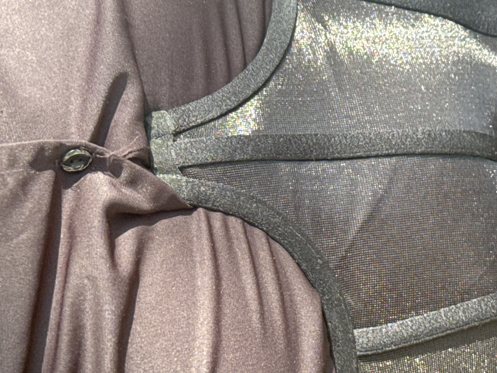 The image is a close-up shot of clothing, specifically a top with a dark, slightly shimmering fabric and a dark grey, structured undergarment with a metallic sheen. The top is ruched, with a button visible near the center. The lighting creates shadows and highlights on the fabric, emphasizing its texture and folds. The scene is not identifiable as being in Perazića Do, Montenegro.