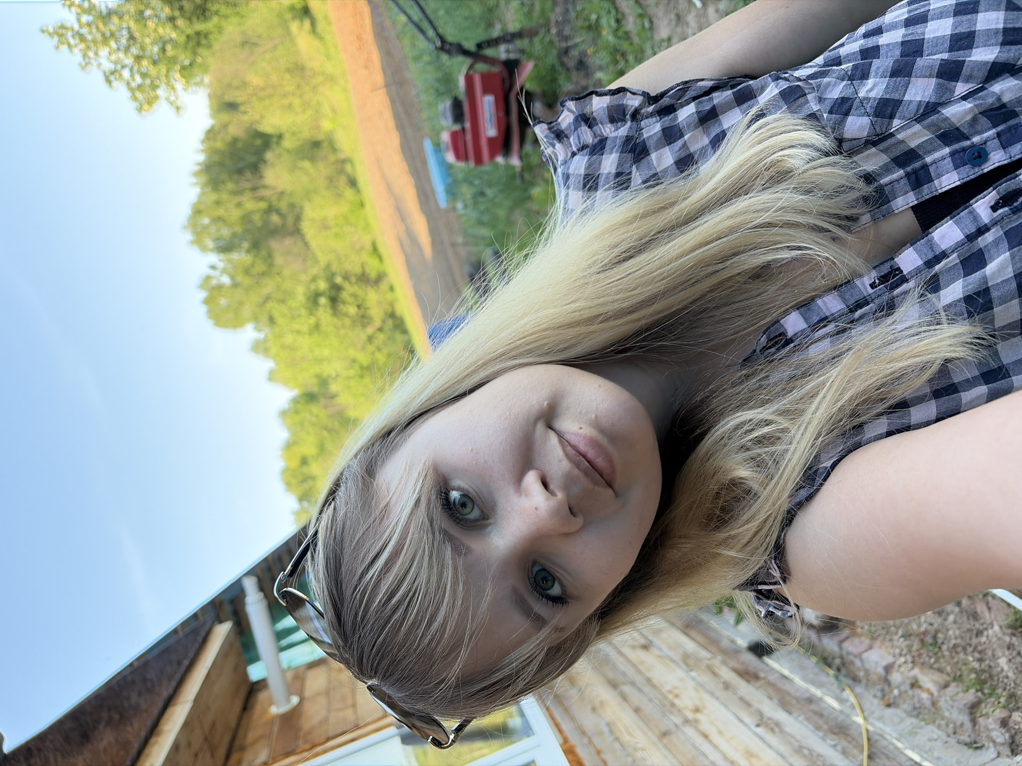 A woman with blonde hair and green eyes is in the foreground, wearing a blue and white checkered shirt. She is standing on a wooden deck and is looking directly at the camera. A pair of sunglasses rests on her head. In the background, there is a field with trees and a small red machine. The sky is light blue. The location is Чумна, Russia, but it is not possible to give more specific details.