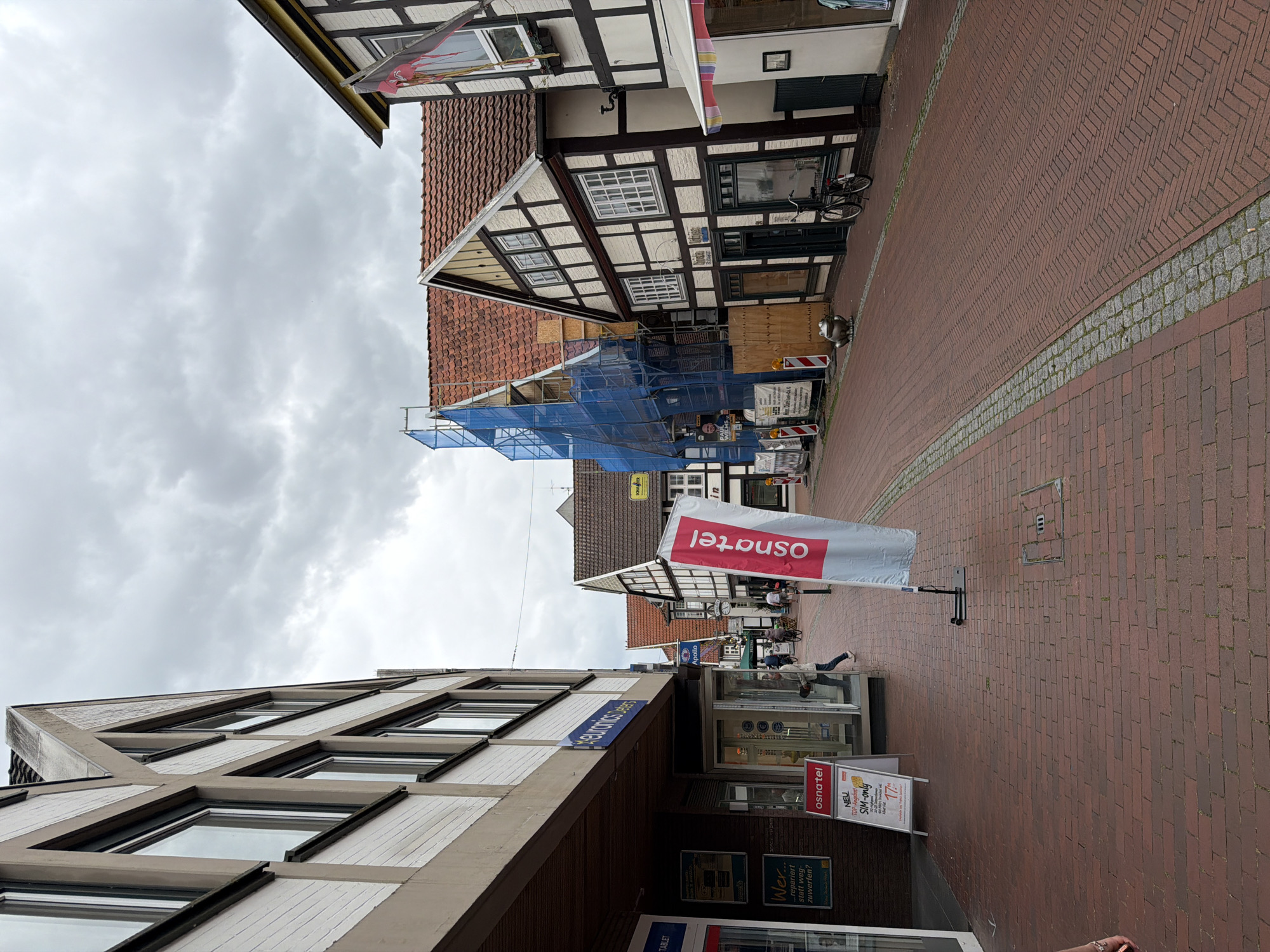 The image depicts a street scene in a European city, likely Germany, based on the architecture. The street is paved with red bricks and a section of cobblestones. Buildings line both sides of the street. On the left, a modern building with white brick and beige trim has large windows and an awning. Several signs are visible, including one for "euronics Deters" and a red and white sign for "osnatel". Further down the street, more traditional half-timbered buildings are present. One building is under scaffolding and covered in blue netting, suggesting renovation. A red flag with an emblem is displayed on a pole in front of a half-timbered building. People are walking on the sidewalk, and a bicycle is parked near a building. The sky is overcast with gray clouds.