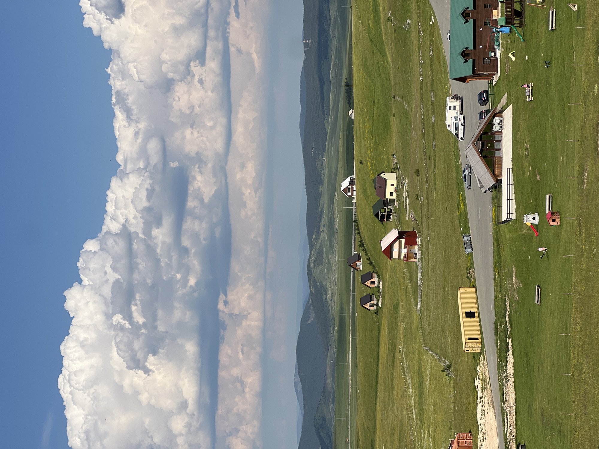 This is an aerial, eye-level, medium shot of a rural landscape in Virak, Montenegro. The image is dominated by a vast, rolling green field under a bright blue sky with large, fluffy white clouds. In the foreground, several structures are visible: a long, yellow shipping container, a brown building with a green roof, and a smaller wooden building with a pitched roof. There are also some recreational structures, including a slide and a playhouse. A few cars and an RV are parked in a paved area near the buildings. In the middle ground, there are several small, wooden houses with dark roofs scattered across the field. The background consists of a series of rolling, green hills and mountains stretching to the horizon. The overall scene depicts a serene, semi-developed rural area.
