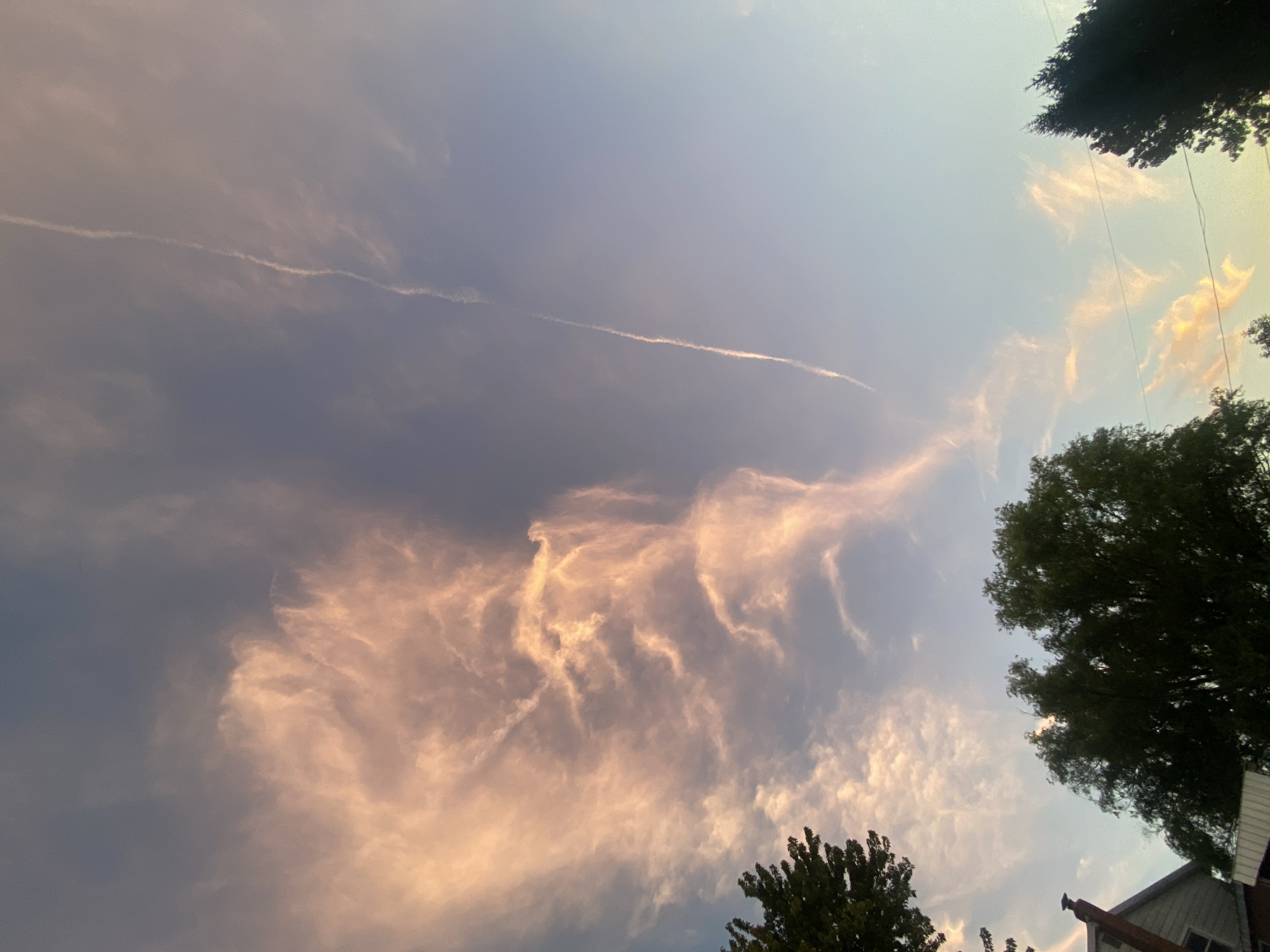 A low-angle, full shot captures a sky filled with wispy, pink-tinged clouds. A long, thin contrail slices diagonally across the upper right portion of the frame. In the foreground, the dark silhouettes of trees and a portion of a house are visible against the bright sky. The house has a brick chimney and white siding. The overall impression is of an evening sky with dramatic cloud formations.