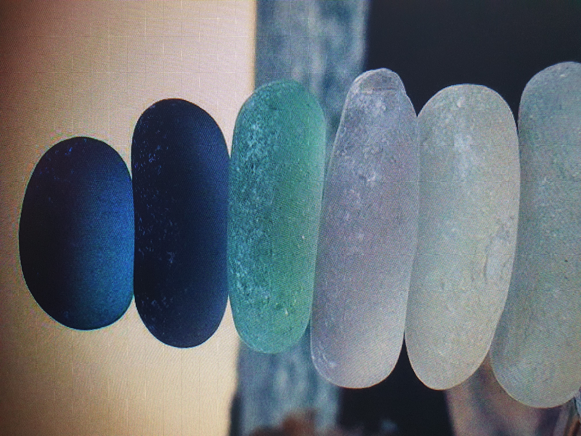 A vertical stack of six smooth, rounded sea glass pieces is centrally framed against a blurred background of water and sky. The sea glass ranges in color from deep blue at the top, through dark blue, teal, light blue-gray, white, and finally a very pale blue-gray at the bottom. The background suggests an outdoor setting, possibly a beach, with a body of water and a hazy horizon. The image has a slightly pixelated quality, indicating it might be a digital photograph viewed on a screen. There are no people, animals, or man-made structures clearly visible. There is no discernible connection to the city of Tashkent, Uzbekistan, as the scene depicts a natural landscape.