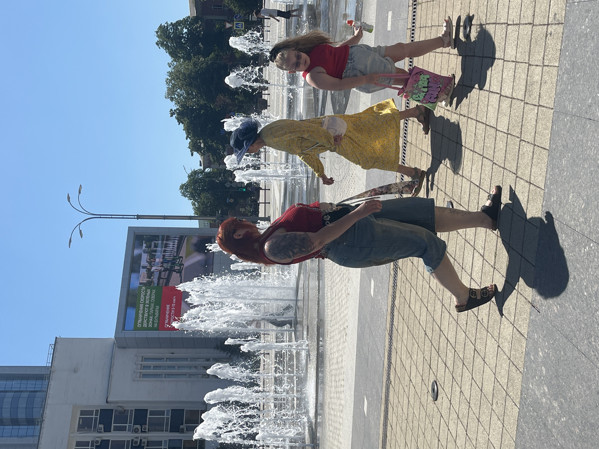 In a bright, sunny outdoor scene, three people are walking near a large fountain. The person in the foreground is a woman with red hair, wearing a red top and denim shorts. She has a large tattoo on her left arm and is looking to her left. Behind her, another woman in a yellow dress and a blue hat walks with her head down. A young girl in a red top and denim shorts is in the background, holding a pink bag and a toy gun. The fountain is spraying water, creating a mist in the air. The ground is paved with light-colored square tiles. In the background, there is a building with large windows and an advertisement that reads, "ОГРАНИЧЕНИЯ СКОРОСТИ ДЕЙСТВУЮТ В ЗЕЛЁНЫХ ЗОНАХ: ПАРКАХ СКВЕРАХ НА БУЛЬВАРАХ ОГРАНИЧЕНИЕ СКОРОСТИ 5-15 км/ч." The overall impression is a pleasant, casual day at a public square with fountains.