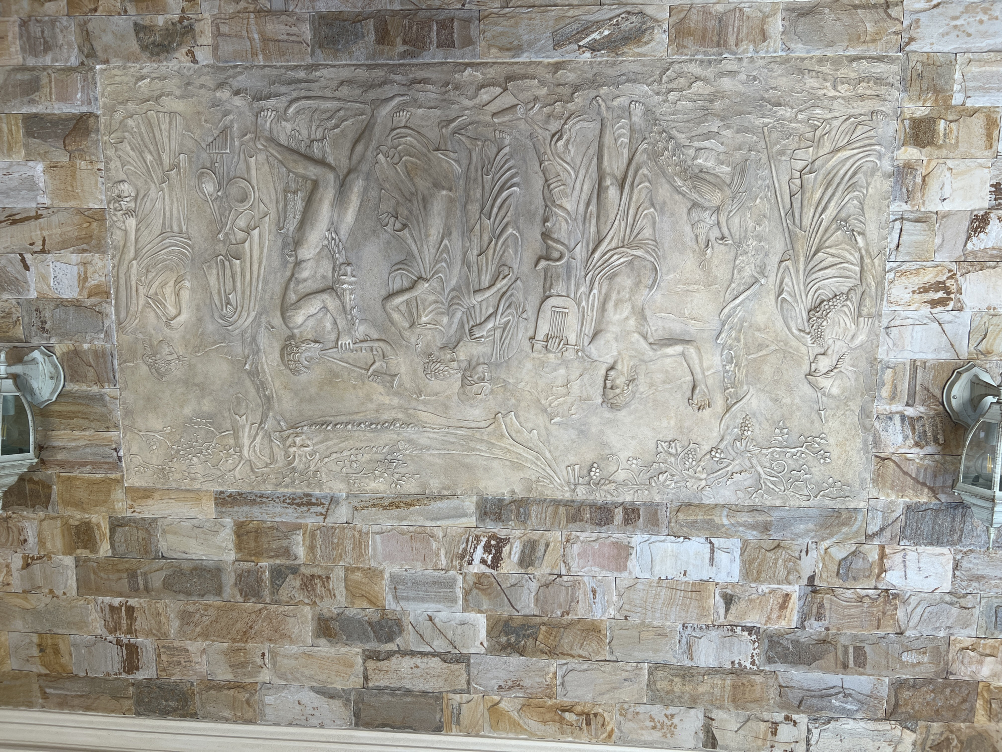 The image displays a detailed bas-relief carving set against a wall constructed of irregular, multi-colored stone blocks. The carving depicts a scene with multiple figures in classical attire, suggesting a historical or mythological theme. On the left, a female figure stands draped in a flowing robe, holding what appears to be a cluster of grapes and a staff, with a peacock at her feet. Next to her, a male figure, also in classical drapery, plays a lyre, with a coiled serpent wrapped around a staff at his side. In the center, two more figures, a man and a woman, stand close together, both clad in robes. To the right, a male figure with a satyr-like head plays a double-flute. Another female figure stands to his right, holding a mask and a laurel wreath. Musical instruments, including a lyre and panpipes, are also visible within the relief. The entire carving is rendered in a light-colored stone, contrasting with the varied earthy tones of the surrounding stone wall. Two wall-mounted lamps with glass casings are visible on either side of the carving, attached to the stone wall. The relief is mounted on the exterior of a building, as suggested by the stone wall construction and the outdoor lighting fixtures. The specific location in Bender, Moldova, is not provided by the visual information alone.