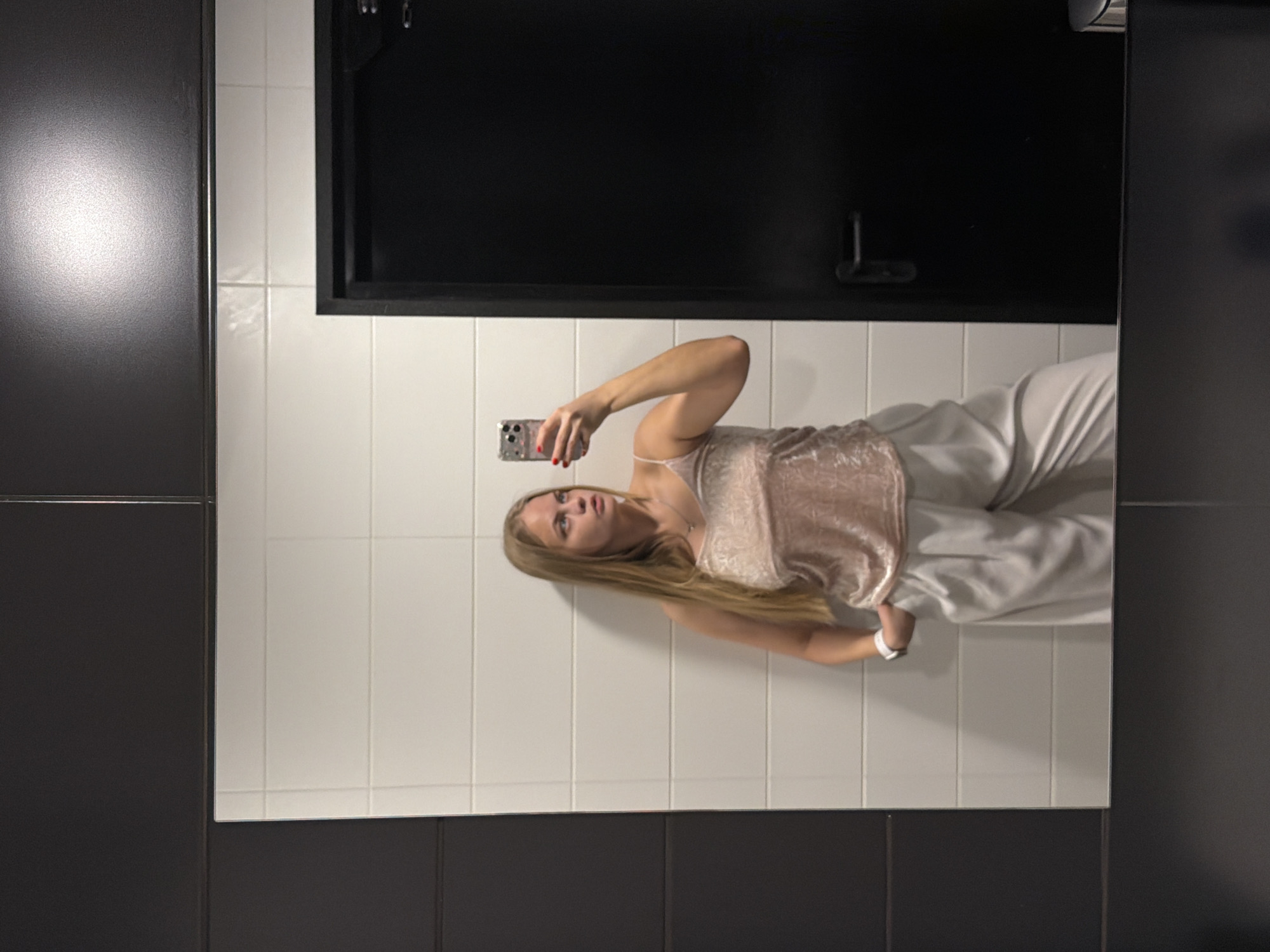 A blond woman in white pants and a pink velvet top is taking a selfie in a bathroom mirror. The bathroom has black and white tiled walls. A black cabinet is mounted above the sink. This photo does not provide enough details to determine the specific location in Izhevsk, Russia.