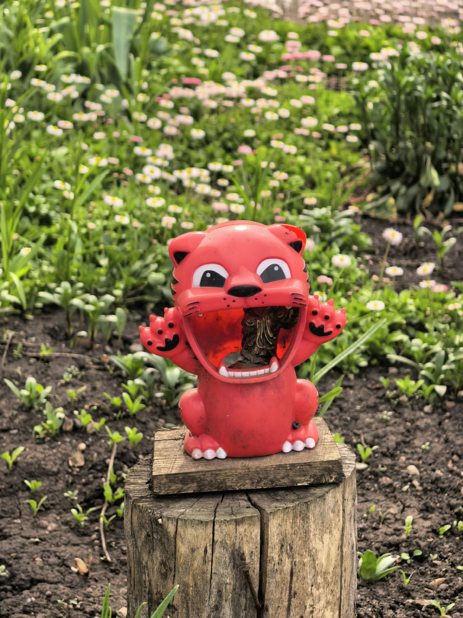A medium shot captures a red, cartoonish cat statue with an open mouth, positioned on a wooden stump in a garden setting. The statue is seated on a small, rectangular piece of wood placed atop the stump. The background is filled with greenery and small white and pink flowers, suggesting a garden or park environment. The surrounding soil is dark, with small green plants emerging from the ground. The photograph was taken on a clear day, but the precise location in Sterlitamak, Russia, is not visually determinable from the given image.