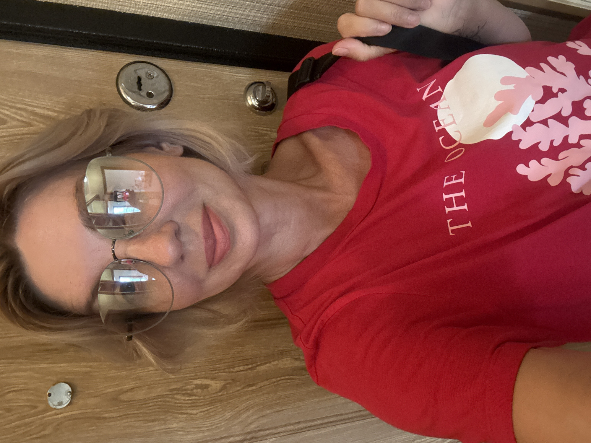 The image shows a woman wearing a red t-shirt with a white and pink design that reads "THE OCEAN." She has blonde hair and is wearing large, round eyeglasses. She is holding a black strap over her shoulder, and her right hand is visible, appearing to adjust the strap. The background consists of a wooden door with two metal fixtures, likely door handles or locks, and a textured fabric surface to the right. The overall setting suggests an indoor environment, possibly a home or hotel room, with the door indicating an entrance. There is no visual information within the image to associate the location with the city of Sochi, Russia.