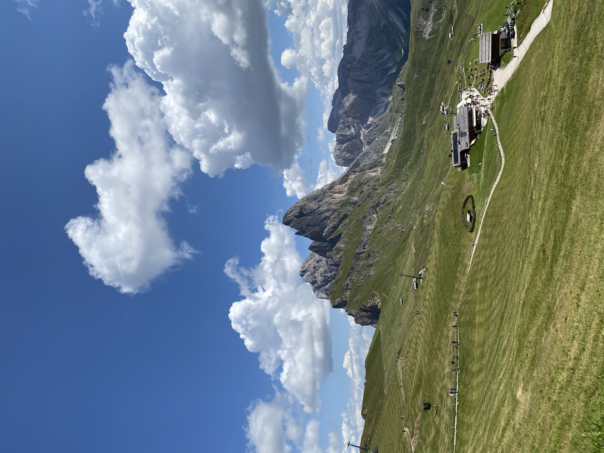 The image depicts a scenic mountain landscape in the Dolomites, likely near Santa Cristina Gherdëina - St. Christina in Gröden - Santa Cristina Valgardena, Italy. The foreground is dominated by rolling green grassy slopes with visible mowing patterns, suggesting well-maintained tourist areas. Several winding dirt paths traverse the landscape, with small groups of people visible walking along them.
In the midground, a cluster of buildings with wooden roofs, resembling a mountain restaurant or lodge complex, is situated on a grassy plateau. Numerous people are gathered around these buildings, some sitting at outdoor tables, others standing or walking. The complex is accessible by a road or wide path, where a few cars are parked. A small, circular fenced area is also visible near the buildings.
Further into the background, majestic, rugged mountain peaks rise sharply against a bright blue sky dotted with fluffy white clouds. The mountains are characterized by rocky, steep slopes, some with patches of green vegetation and others appearing more barren and stony. A ski lift mechanism, consisting of towers and cables, is visible on the left side of the frame, indicating the area's use for both summer and winter activities. The overall scene conveys a sense of natural beauty and outdoor recreation.
