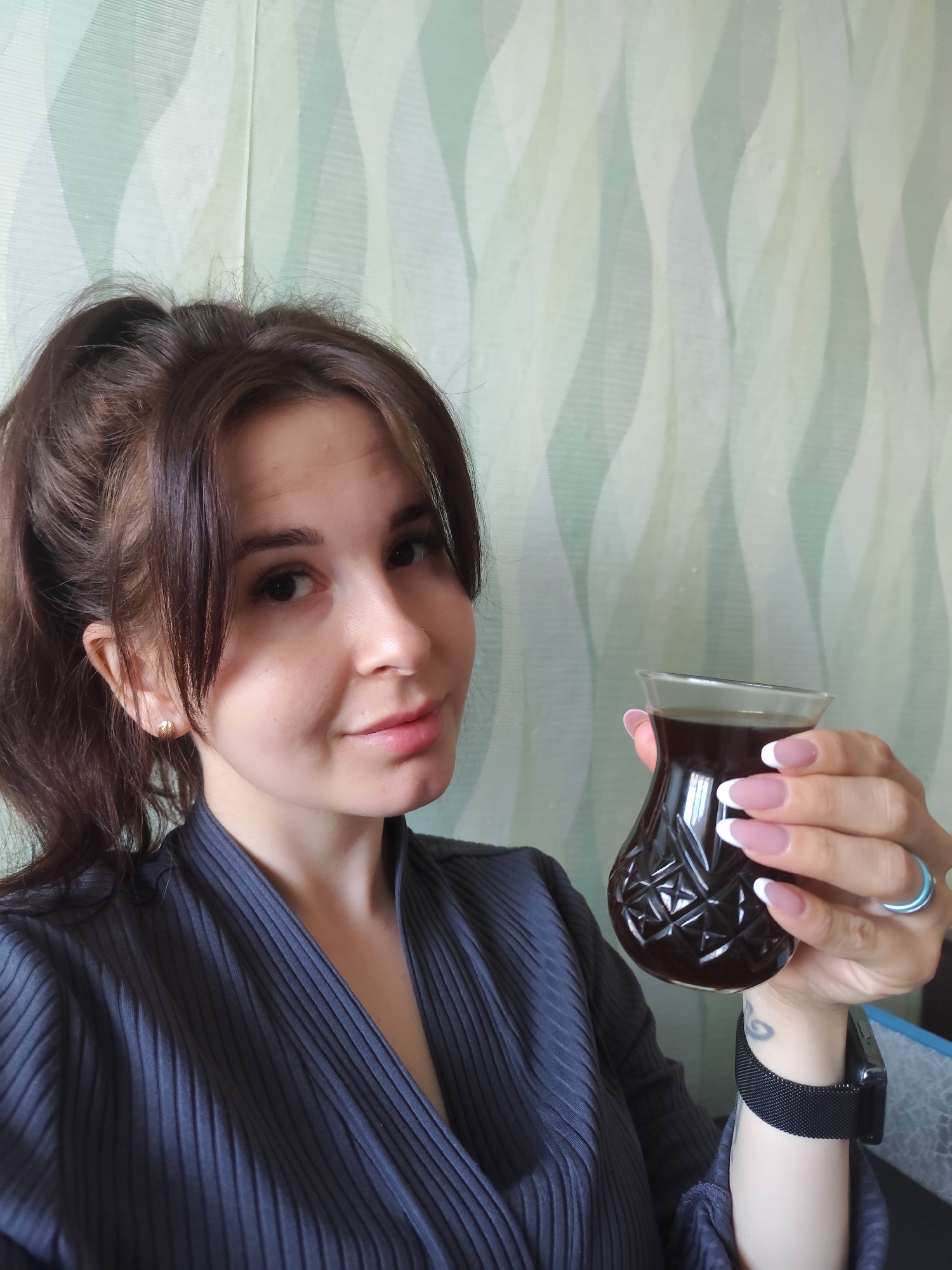 A young woman with brown hair pulled back in a ponytail holds a glass of dark liquid. She has a French manicure and wears a gray ribbed V-neck garment. A black smartwatch adorns her left wrist, and a blue ring is on her right hand. The background is a light green wallpaper with a wavy pattern. The location Фоки, Russia cannot be determined from the image.