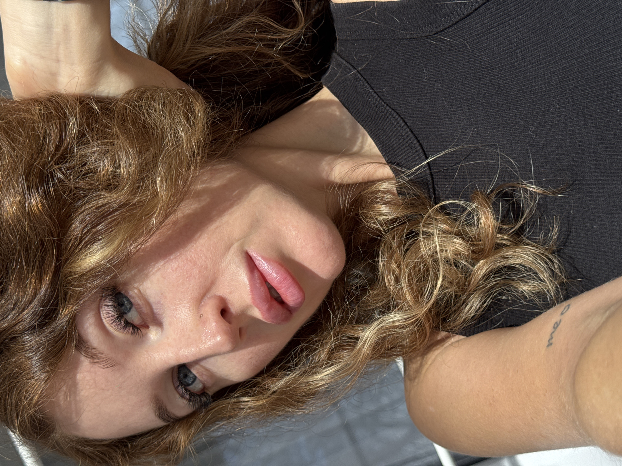 A young woman with wavy, light brown and auburn hair is pictured in a close-up, laid-back pose. The image is oriented vertically, showing her head and upper torso as if she is lying on her side, looking directly at the viewer. Her hair is spread around her head and shoulders. She has striking blue eyes with prominent, dark eyelashes, and a small stud piercing on her left nostril. Her lips are full, pink, and slightly parted.
She is wearing a black ribbed top. Part of her left arm is visible in the upper left of the frame, and her right forearm, bearing a small, cursive tattoo that appears to read "meo," is visible on the right. The lighting is strong and directional, suggesting natural sunlight, creating distinct shadows across her face and body. The background is largely out of focus, indicating a shallow depth of field, with hints of a lighter surface beneath her and possibly a dark background behind her. The overall mood is relaxed and direct. This scene takes place in Chaikovsky, Russia.