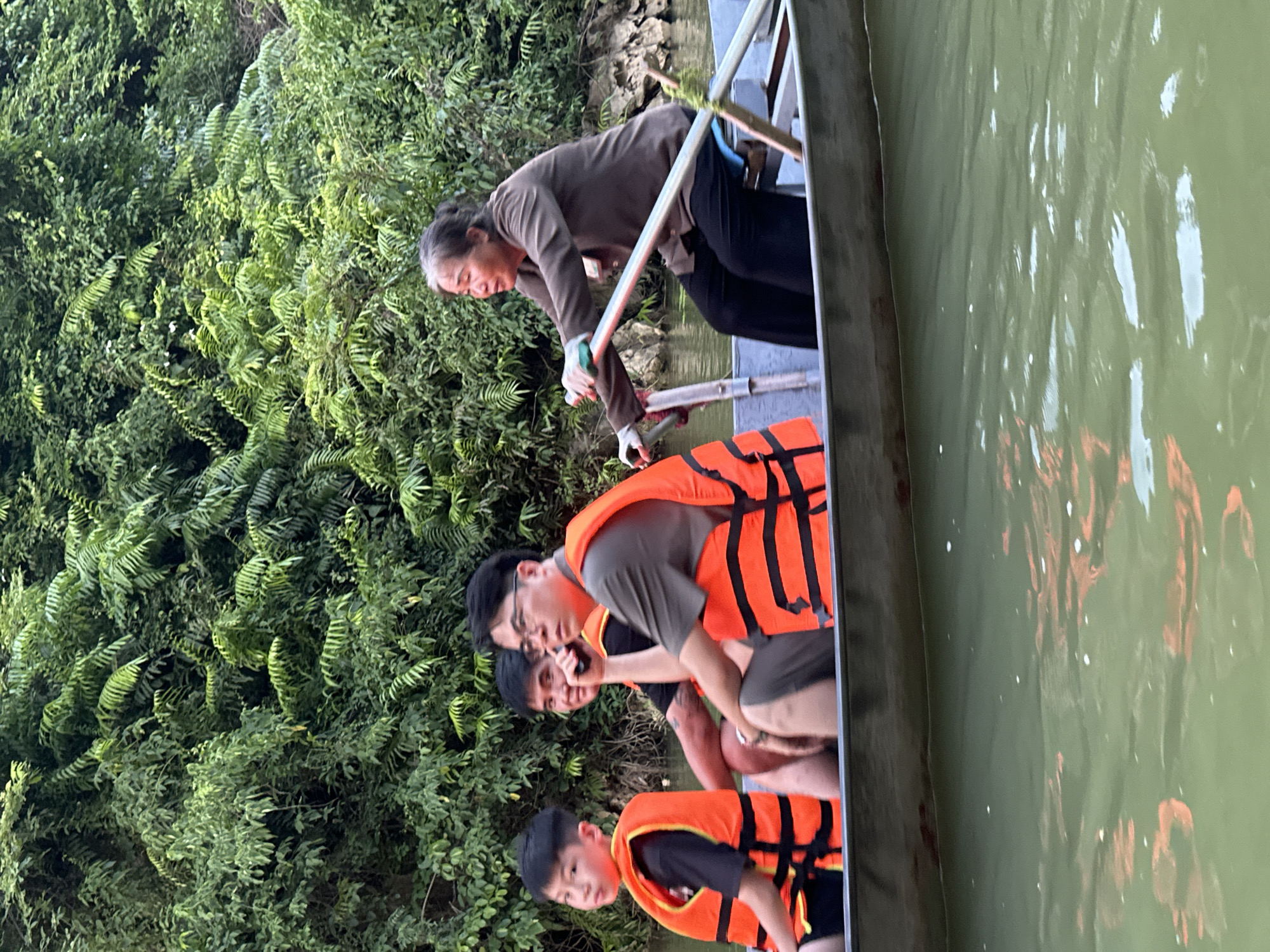 Four individuals are in a rowboat on a river, surrounded by lush green foliage. The person at the rear of the boat, an elderly woman, is rowing with a pair of oars. She is wearing a brown long-sleeved shirt and black pants, with white gloves on her hands. In front of her, from right to left, are a man wearing a grey shirt and black pants, and two younger individuals, one in a black t-shirt and orange life vest, and the other in a black t-shirt and orange life vest. The river appears calm, and the overall scene suggests a tranquil moment on the water. The dense vegetation lining the riverbanks indicates a natural, possibly rural setting, consistent with locations such as Yên Sơn Ward in Vietnam.