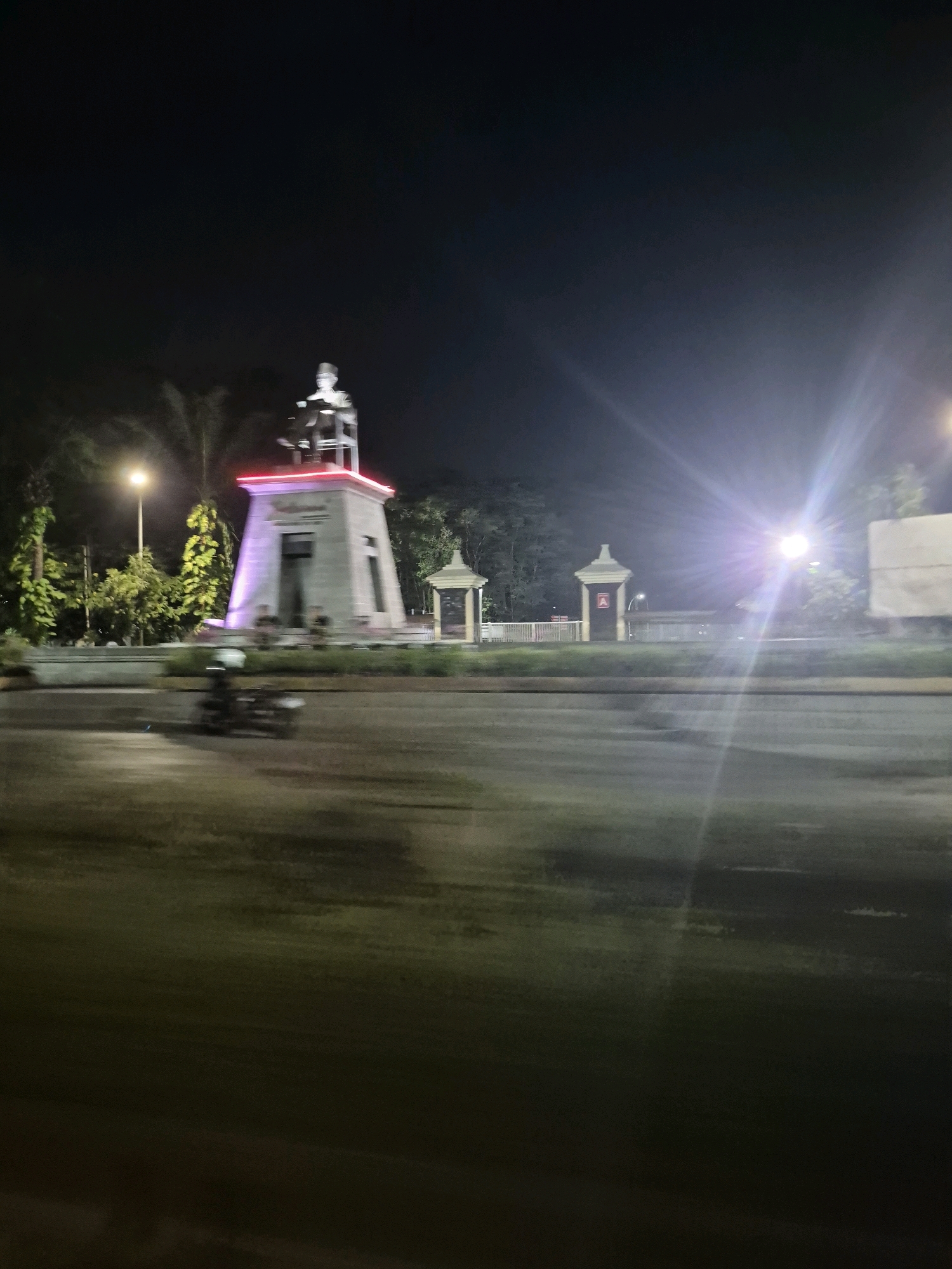 The image captures a nighttime view of the Slamet Riyadi Monument, a historical landmark situated on Jalan Slamet Riyadi in Surakarta, Indonesia. The monument, a white, tiered structure topped with a statue, is illuminated by purple and red lights, creating a striking contrast against the dark sky. In the foreground, a motorcycle with a rider is visible, captured in motion, suggesting a road or intersection environment. The street lights and lens flare from a bright light source add to the nocturnal ambiance. The surrounding area appears to be an urban park or public space with trees and other structures that are partially visible. The overall impression is a well-lit monument in a city setting at night.