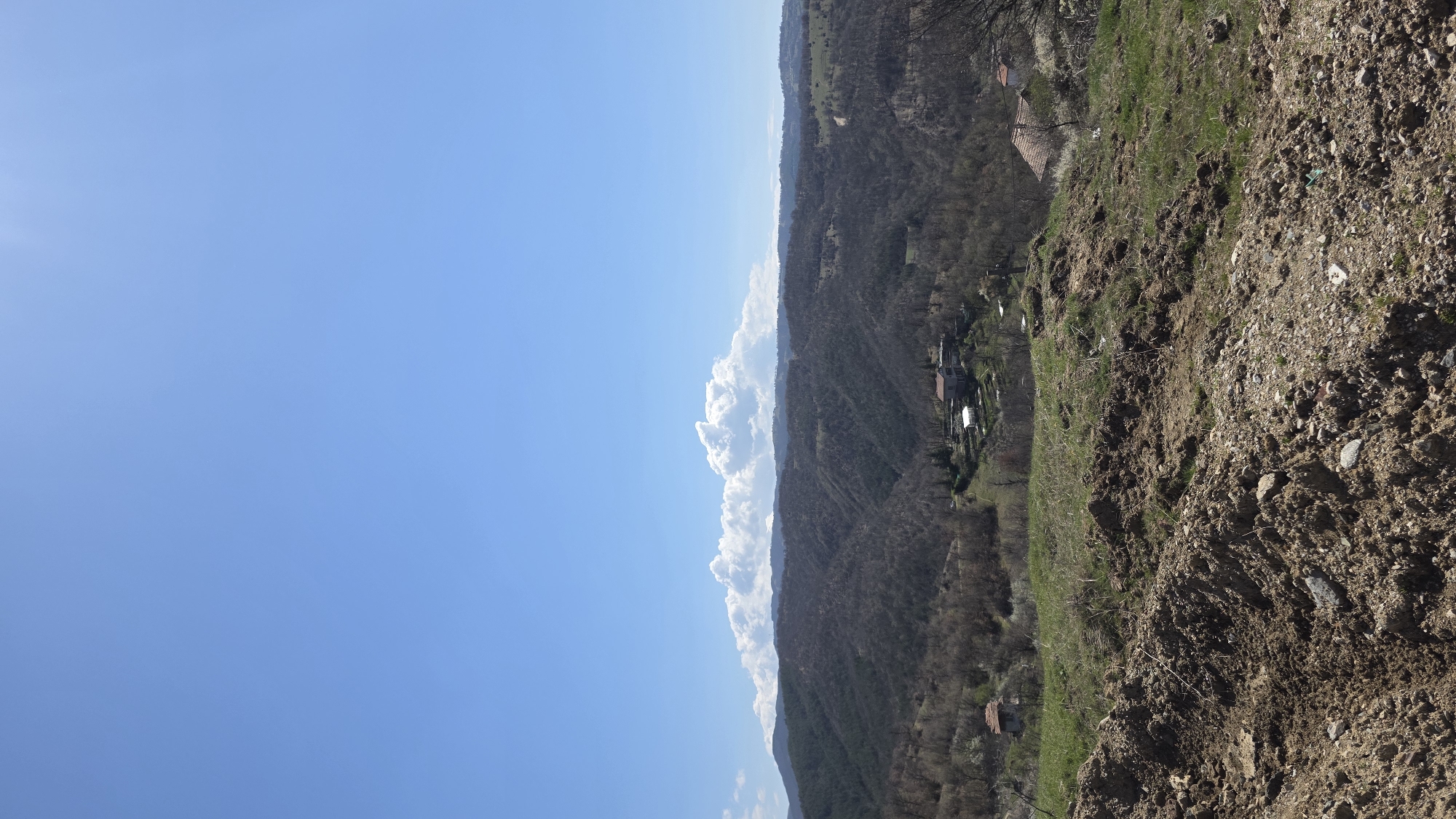 The image presents a view from an elevated position in Marulevo, Bulgaria, looking towards a valley. The foreground consists of rocky, uneven terrain transitioning into a hillside covered with sparse vegetation. In the middle ground, a deep valley is visible, densely populated with trees and a few scattered buildings and houses. The sky is clear blue, with white clouds lining the horizon, partially obscuring distant hills or mountains. No people are visible in the frame.