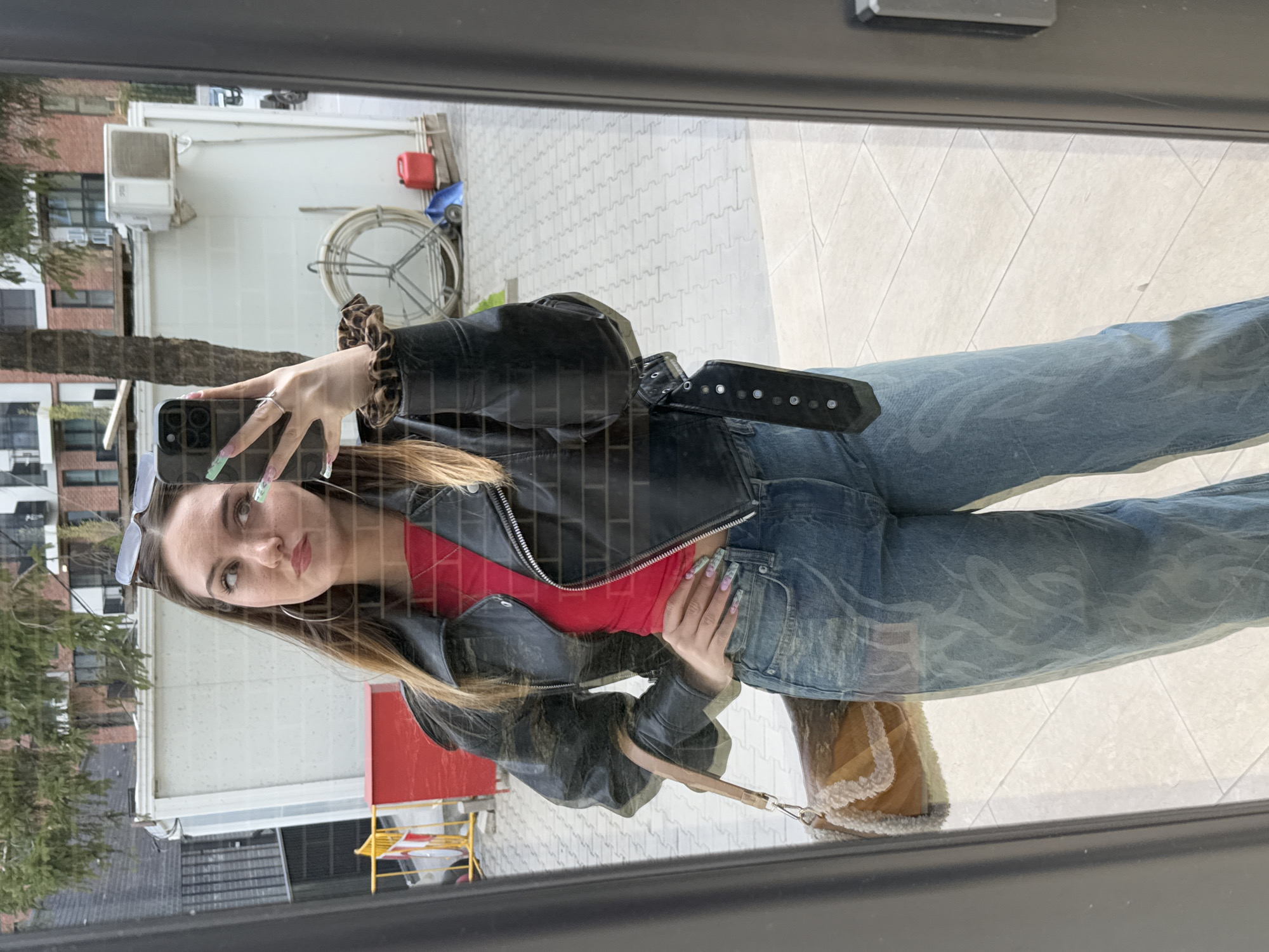 A young woman is taking a selfie in what appears to be an outdoor setting. The setting is a city, specifically Sofia, Bulgaria, based on the architecture visible in the background. The woman is wearing a red top under a black leather jacket, light-colored jeans with a pattern, and is carrying a small brown handbag. She is holding a smartphone with her left hand, visible due to the reflection in the glass she is taking the selfie in, and has long, patterned fingernails. Her hair is light brown, and she is wearing sunglasses on her head. The background reveals a building with an air conditioning unit, a paved area, some greenery, and street furniture. The reflection captures the overall setting, suggesting a bright, sunny day.