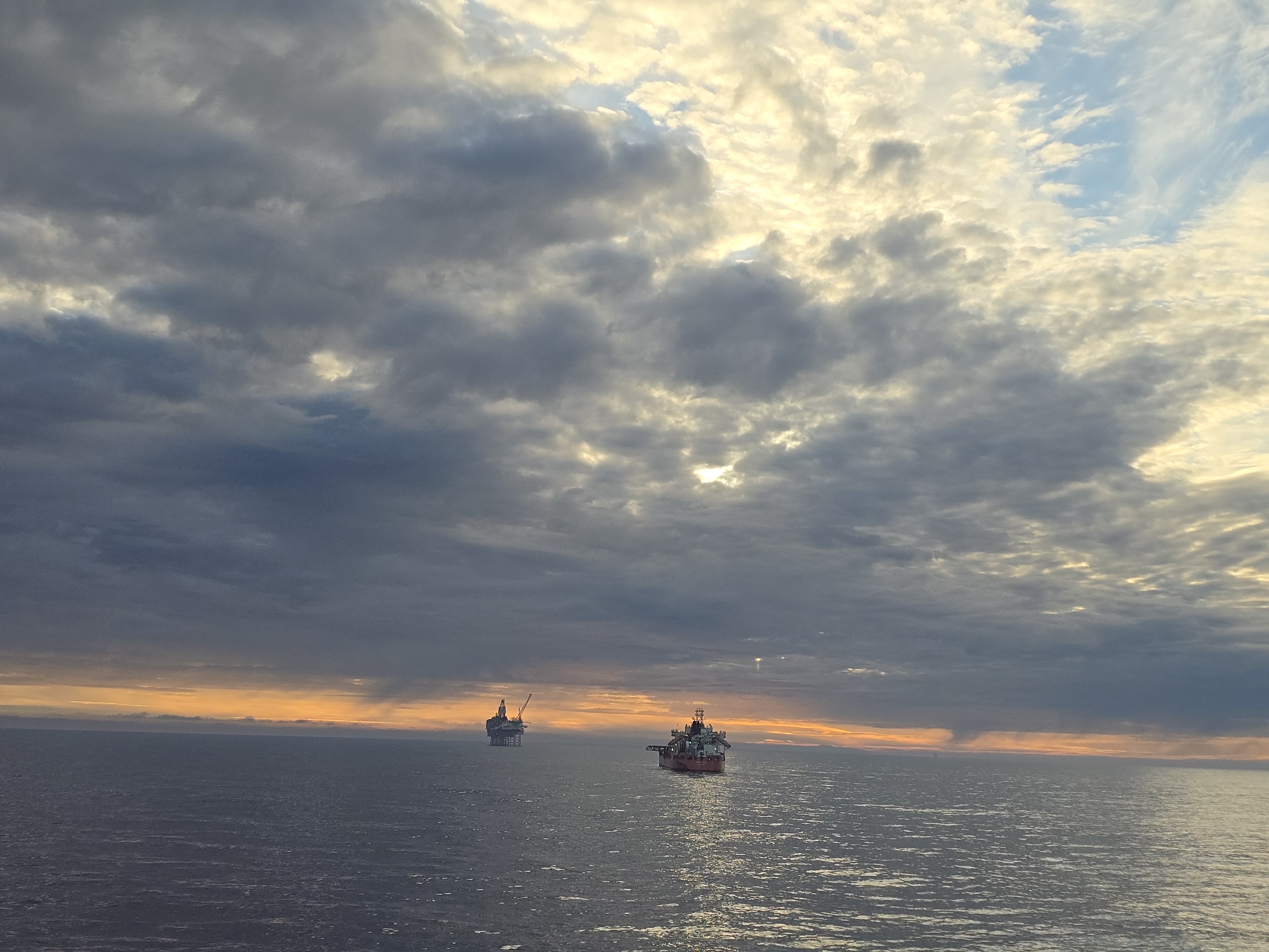 The image depicts a seascape under a dramatic sky dominated by heavy, textured clouds. A mix of grey and white clouds are illuminated by the setting sun, which casts a warm, golden glow across the horizon and parts of the cloud cover. The sea is relatively calm with small ripples reflecting the light. In the distance, two structures are visible on the water: one appears to be an oil rig, and the other is a ship. The location is an open sea environment; the city is unknown.