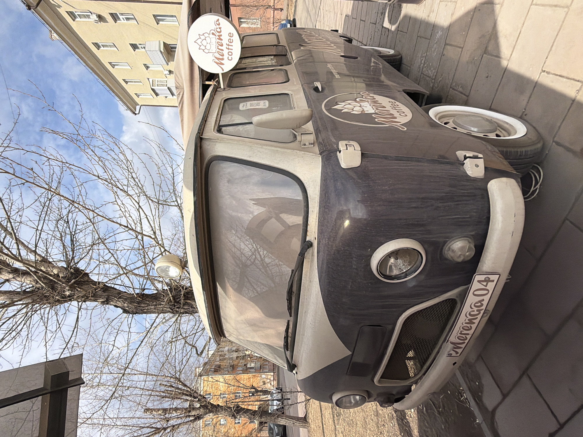 The image shows a vintage van, likely a Volkswagen Type 2, modified into a mobile coffee stand. The van is parked on a brick-paved area, presumably a sidewalk or pedestrian zone. The van is two-toned, with a light top and a darker blue/gray bottom. The business name, "Merenca coffee," is prominently displayed on the side and a round sign on the roof. The license plate reads "Merenca 04." The van is positioned in a street setting, with buildings and trees visible in the background. The location appears to be Krasnoyarsk, Russia, based on the name on the license plate. The trees are bare, suggesting it is either late fall or early spring. There are no people visible in the immediate foreground.