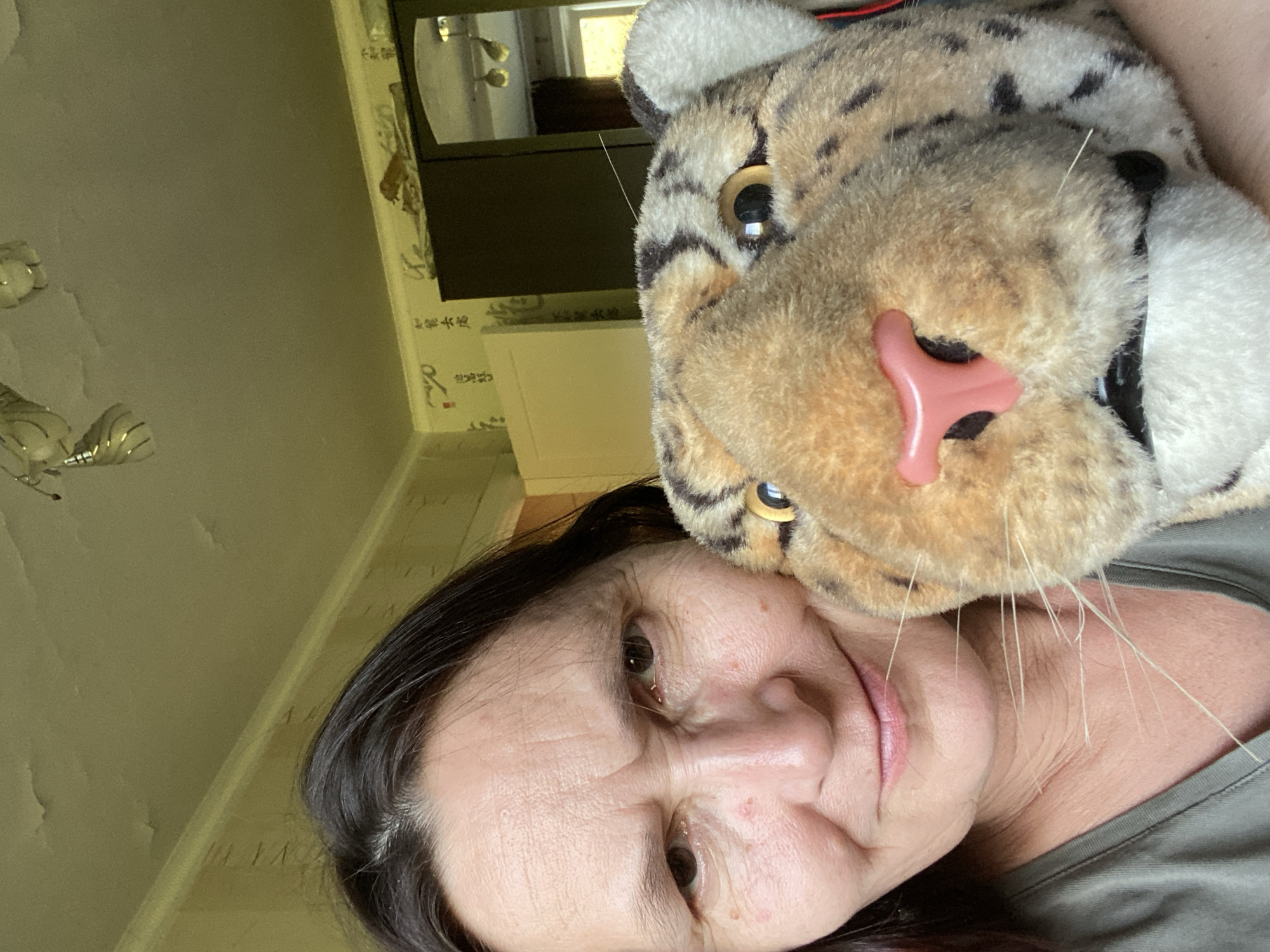 The image is a close-up selfie of a woman and a stuffed toy animal, likely a leopard, taken indoors in what appears to be a residential setting. The woman, with dark hair and fair skin, is on the left side of the frame, her face angled towards the camera with a slightly furrowed brow. Her facial features include brown eyes, a relatively prominent nose, and a closed-mouth smile. She is wearing a green t-shirt. The stuffed animal is positioned to her right, with its large head filling a significant portion of the right side of the image. Its face features yellow eyes with black pupils, a pink nose, and grey and black spots on its fur. Several long, white whiskers extend from the toy.
The background suggests an indoor environment. The ceiling is visible at the top, appearing to have some peeling paint or discoloration. A light fixture with multiple bulbs is attached to the ceiling. Below the ceiling, a patterned wallpaper is visible on the wall, featuring what looks like Asian calligraphy or designs. To the right, a dark-colored wardrobe or closet with a mirrored door is partially visible. A white cabinet or door is also visible to the left of the wardrobe. The lighting in the room seems to be artificial, casting a warm glow. The overall impression is of a casual, personal moment captured in a home. There is no indication of the specific location within Novokuznetsk, Russia, beyond it being an indoor residential space.