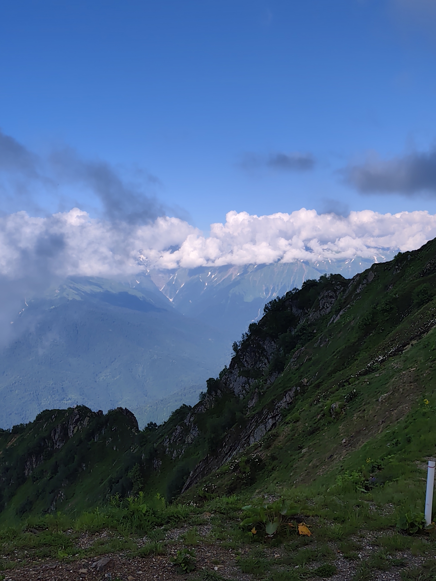 This is a vertical, eye-level, medium shot of a mountain landscape. The sky is a vibrant blue with scattered white and gray clouds. In the middle ground, layers of lush green mountains recede into the distance, some with patches of snow and rocky peaks. A prominent ridge of dark green, rocky mountains covered in trees runs diagonally from the bottom left towards the center of the image. The foreground consists of a rocky, grassy slope. A white pole is visible on the right side of the image. The scene appears to be in the city of Estosadok, Russia, likely in the Caucasus Mountains.