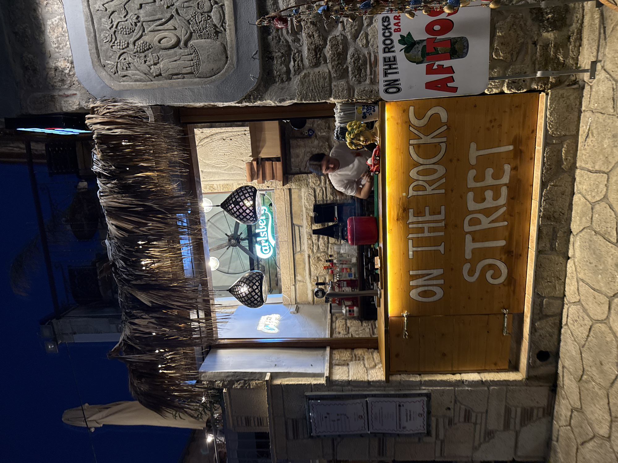 The image depicts a street-level bar in Kassandra Municipal Unit, Greece, during nighttime. The bar, named "ON THE ROCKS STREET BAR AFITOS", features a wooden counter illuminated by warm yellow lights. Behind the counter, a female bartender with dark hair pulled back is visible, wearing a white t-shirt, and appears to be preparing drinks. Various bottles of alcohol and bar tools are arranged on the counter. A neon sign for "Carlsberg" is prominently displayed in the background. The bar is covered by a thatched roof, adding a rustic touch. To the right of the bar, a sign with the bar's name and a picture of a cocktail is displayed, along with decorative elements hanging from a string. The establishment is situated on a stone-paved street, with stone buildings surrounding it. The overall atmosphere is that of a lively nighttime establishment.