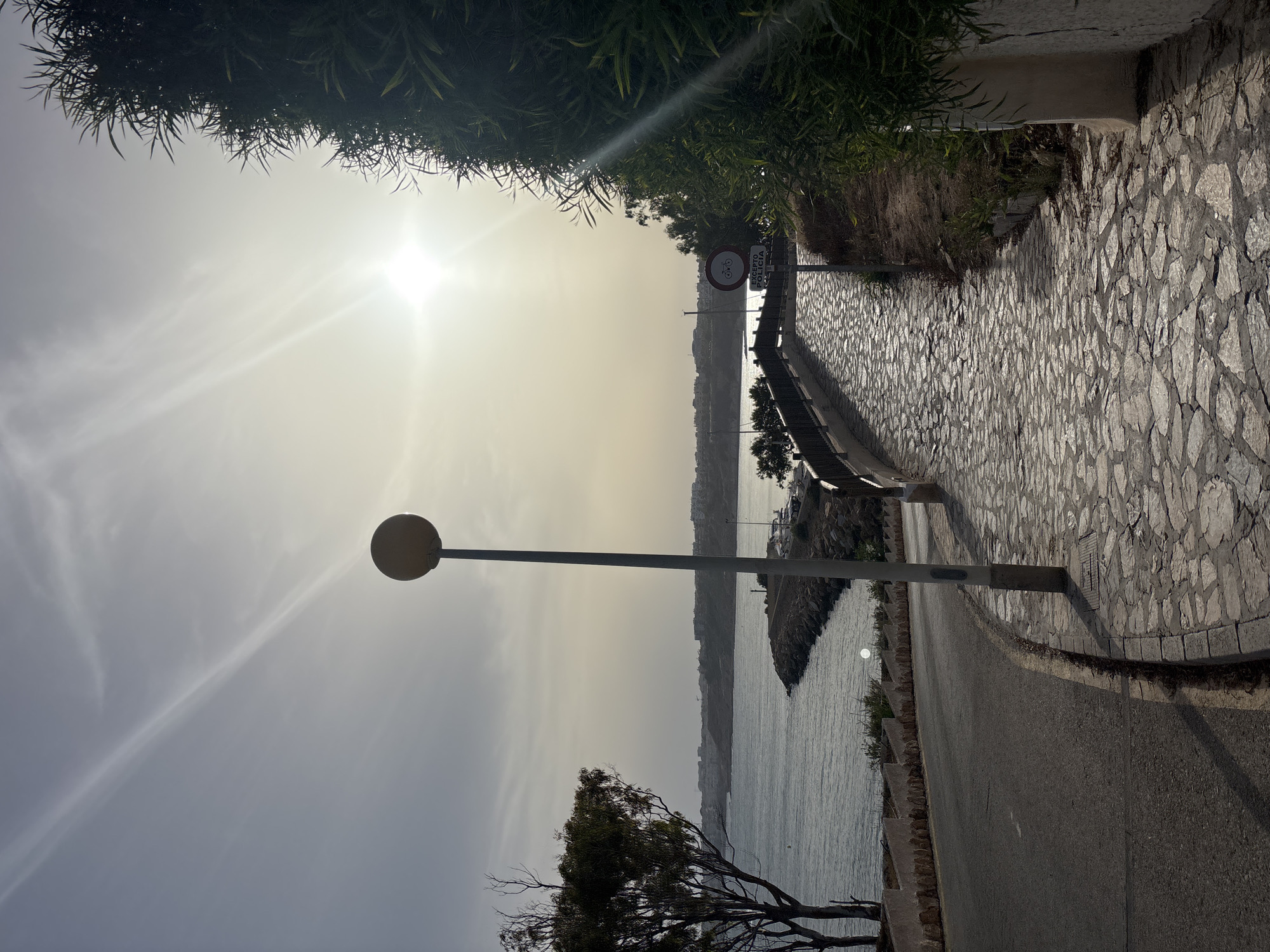The image depicts a paved pathway with irregular stone slabs winding upwards, flanked by lush green foliage on the right and a gentle slope leading to a road on the left. A lamppost with a circular yellow light fixture stands on the left side of the pathway. In the distance, the sea stretches out, with a rocky breakwater extending into it. A town or city skyline is visible along the coastline. A traffic sign prohibiting bicycles is placed near the pathway. The sun is bright, casting lens flares and rays across the sky, indicating it is likely late afternoon or early morning. The environment appears to be a coastal urban area within Orihuela, Spain, possibly a promenade or a park area overlooking the sea. There are no people visible in the image.