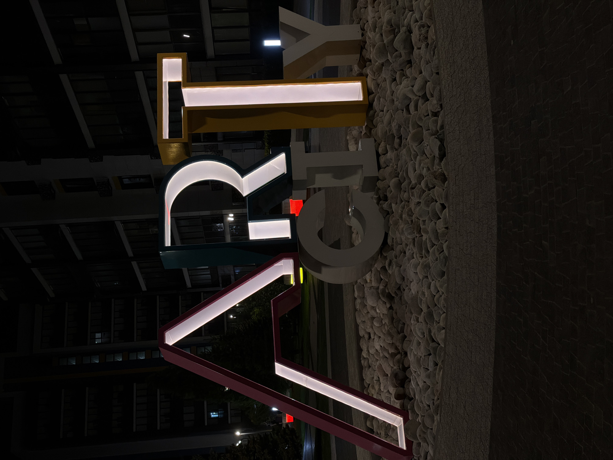 The image depicts the illuminated letters "ART CITY" in Kazan, Russia, at night. The letters are large, three-dimensional, and each painted a different color. The "A" is red, the "R" is teal, the "T" is yellow, and the "CITY" is white. The letters are backlit with white light. The letters are positioned against a background consisting of a wall covered in small, rounded stones. Behind the letters and to the left of the frame is the facade of a modern building with multiple stories. Streetlights are visible in the background. The scene is generally dark, with the illuminated letters providing the main source of light.