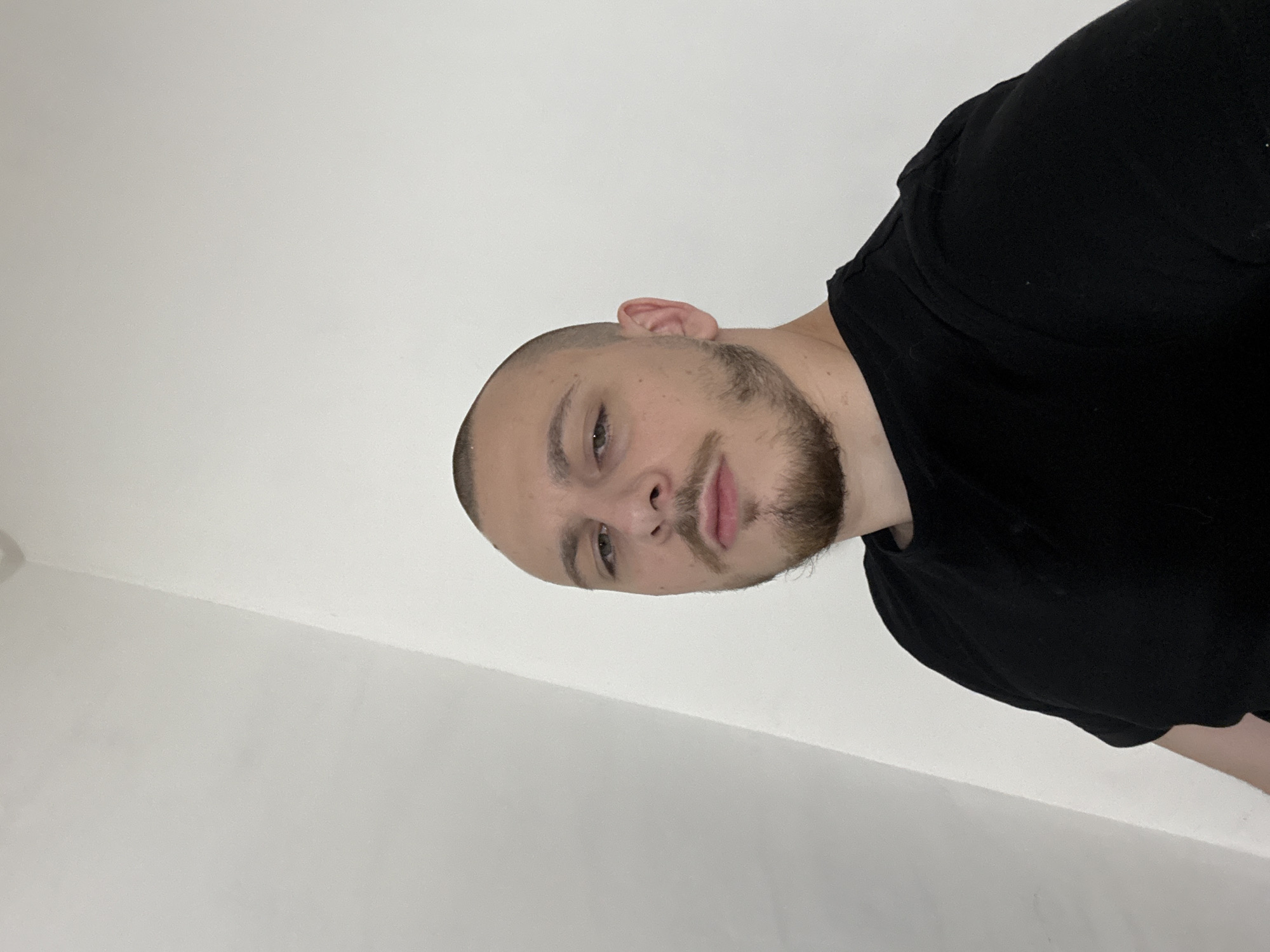 A man with a closely shaved head and a light beard and mustache is pictured. He is wearing a black t-shirt. The background appears to be a plain white wall. The location of the photo within the city of Stara Zagora, Bulgaria is not discernible from the image.
