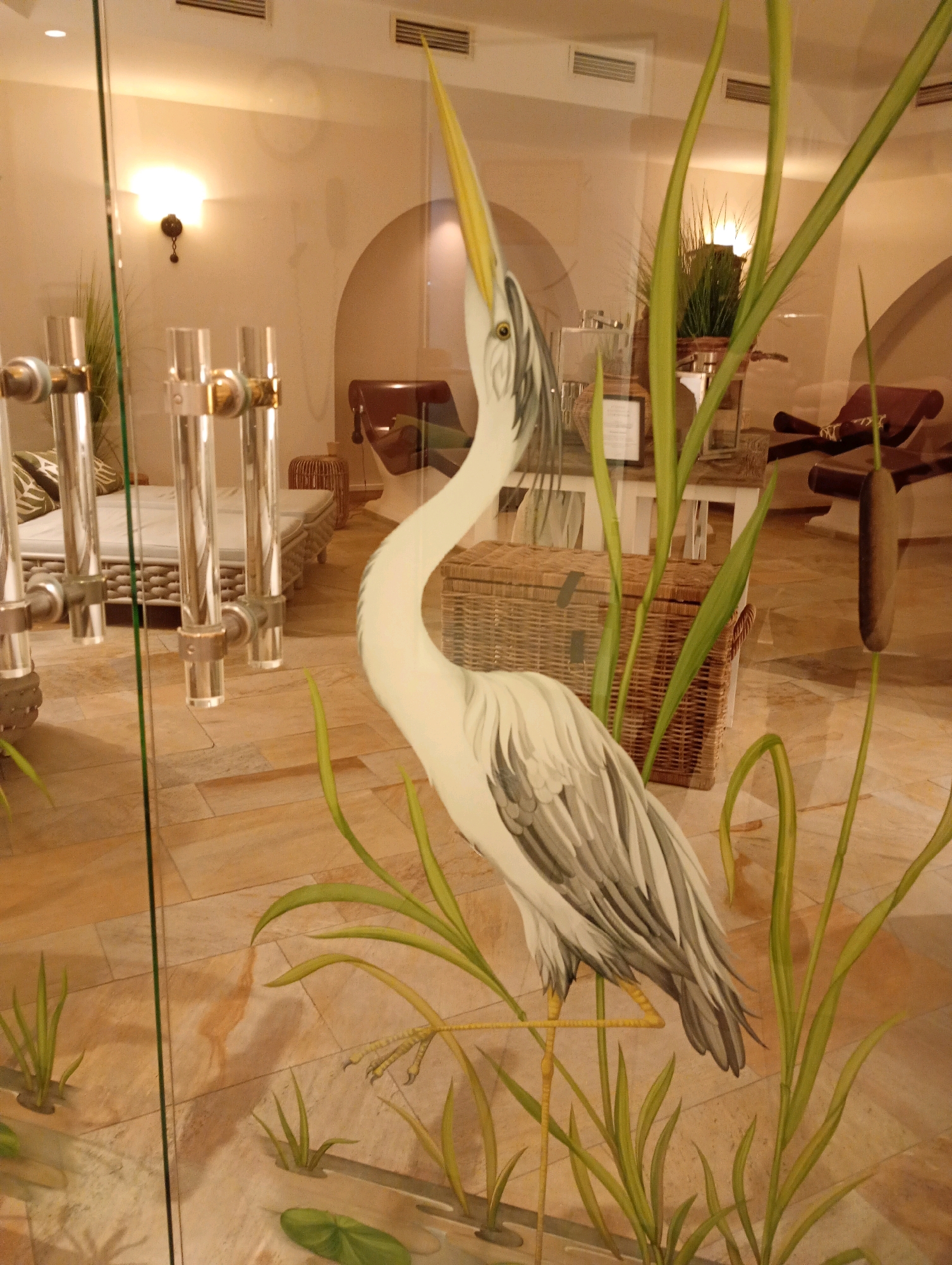 This is an image taken from inside a building, looking out through a glass partition. The partition is decorated with a painted depiction of a heron standing amidst reeds and water plants. In the background, through the glass, is an indoor spa or wellness area. The area features beige tiled flooring, and what appear to be spa beds with light-colored bedding. In the mid-ground, there is a woven basket and furniture that suggests a relaxation space. Lights are visible on the wall, emitting a warm glow. The scene is well-lit, with the overall ambiance suggesting a tranquil and luxurious environment. The location is identified as Bad Sachsa, Germany.