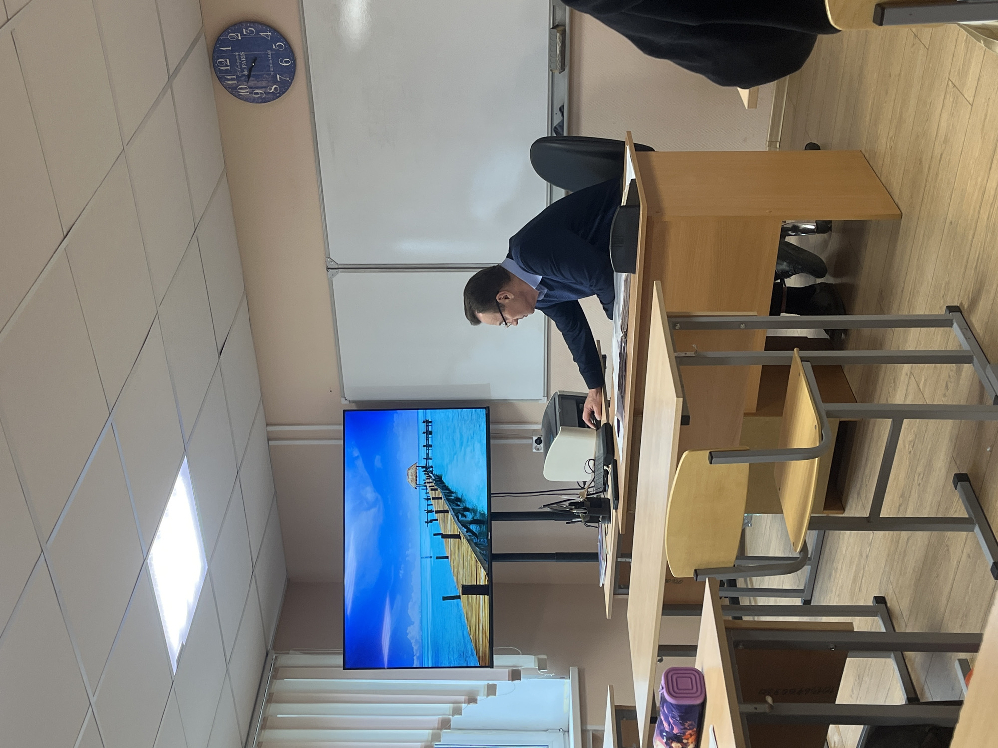 A classroom in Saint Petersburg, Russia, is shown. A middle-aged man in a dark suit and glasses stands at a teacher's desk, looking at a document camera. A large flat screen TV displays a beach scene. Rows of empty student desks with wooden chairs fill the foreground. A clock hangs on the wall.
