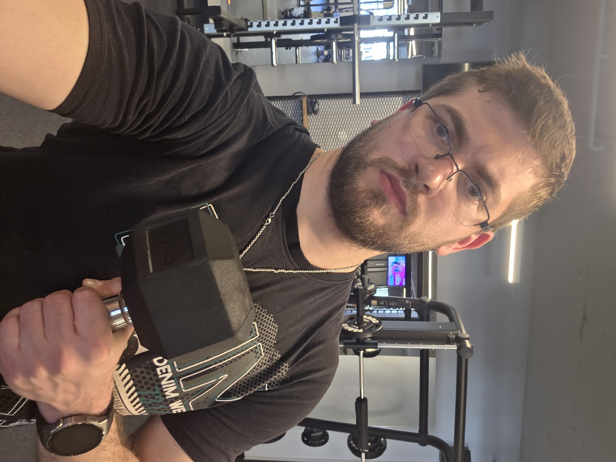 A man with brown hair, glasses, and a beard is holding a dumbbell in a gym. He is wearing a black t-shirt and a silver chain. A smartwatch is visible on his left wrist. The gym is equipped with various weight machines. The location cannot be verified as Plovdiv, Bulgaria without additional information.