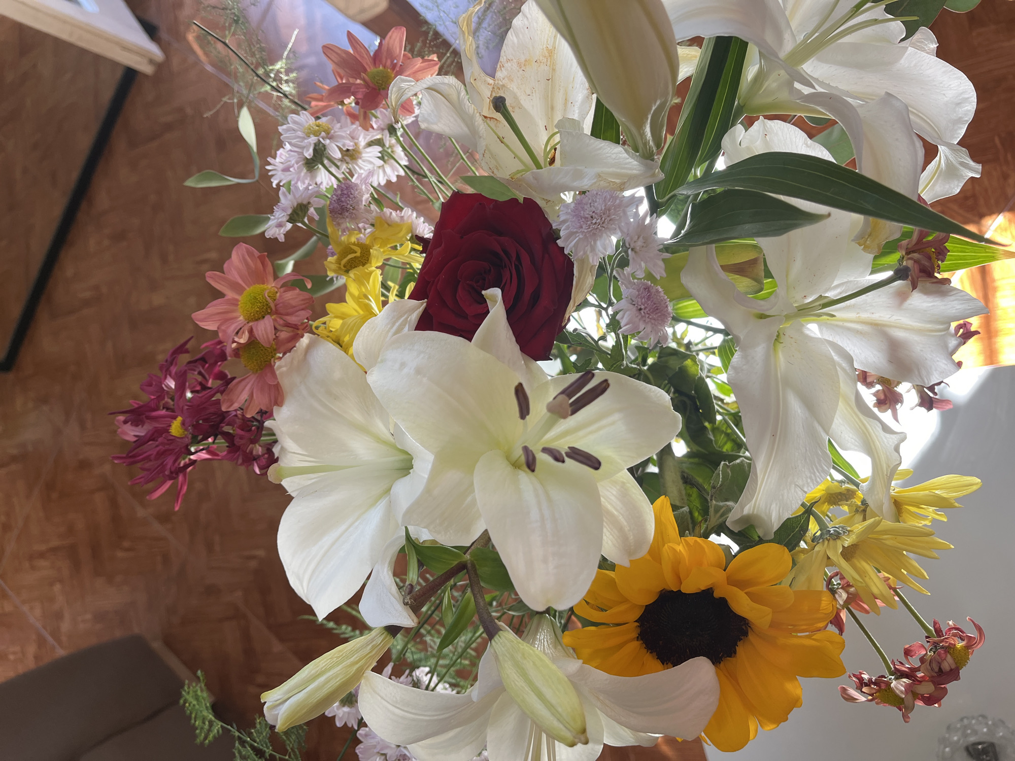 A bouquet of mixed flowers is displayed. White lilies, a single red rose, a sunflower, and other smaller blooms in hues of pink, peach, yellow, and purple are visible. The flowers are densely arranged, with varying textures and shapes, suggesting a natural and slightly wild composition. The background is blurred, hinting at an interior setting. Sunlight streams into the scene, illuminating the flowers. The location is unknown.