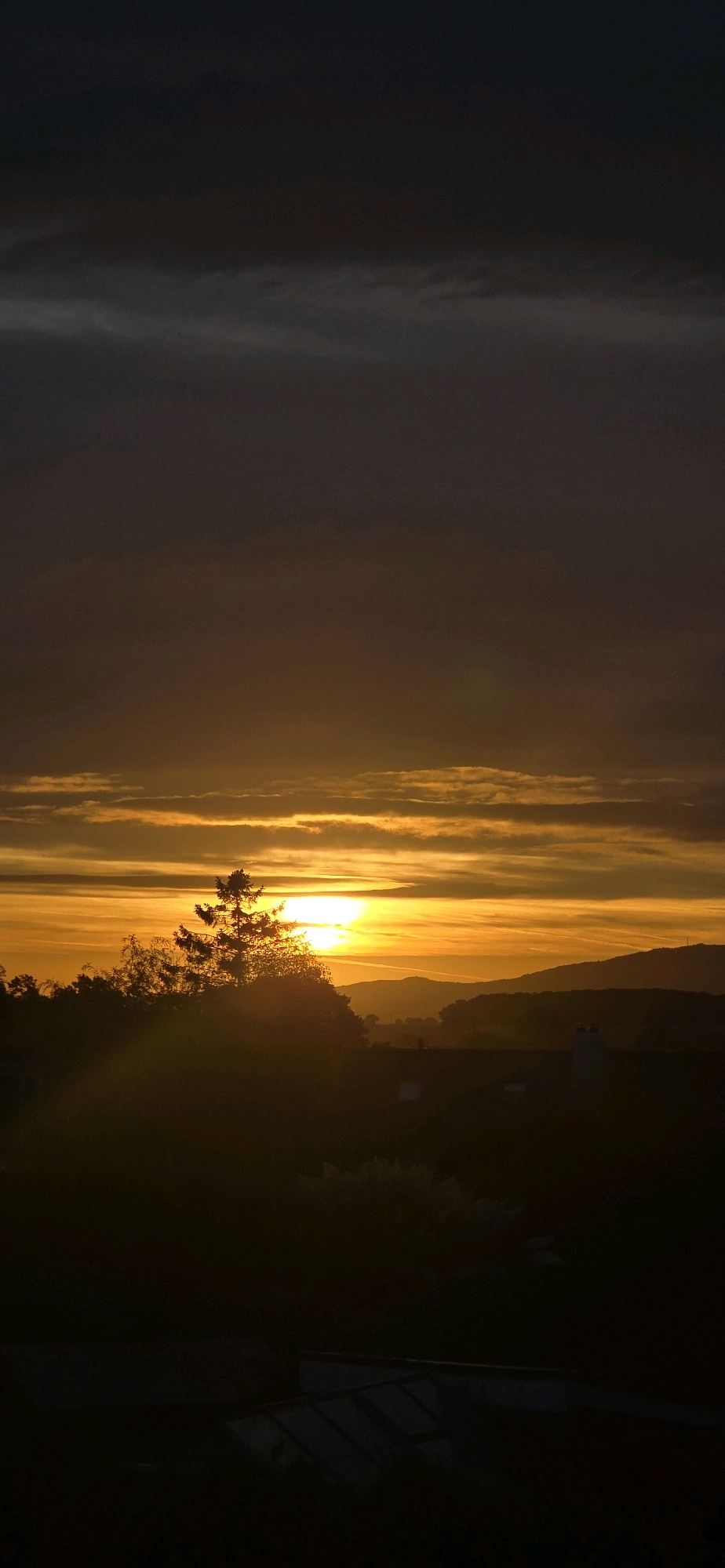 A vertical, full-frame photograph captures a sunset over the town of Bayston Hill, United Kingdom. The sky is dominated by horizontal bands of color, with the sun, a bright, intense yellow orb, positioned just above the horizon, slightly left of the frame's center. Dark, silhouetted trees and foliage occupy the lower third of the image, partially obscuring distant rolling hills. The foreground is very dark, revealing only the faint outlines of structures, possibly rooftops or greenhouses. The clouds in the sky are layered, ranging in color from deep grey and indigo at the top to vibrant oranges and yellows closer to the sun, indicating the sun's descent. The overall lighting is dramatic and atmospheric, characteristic of twilight.