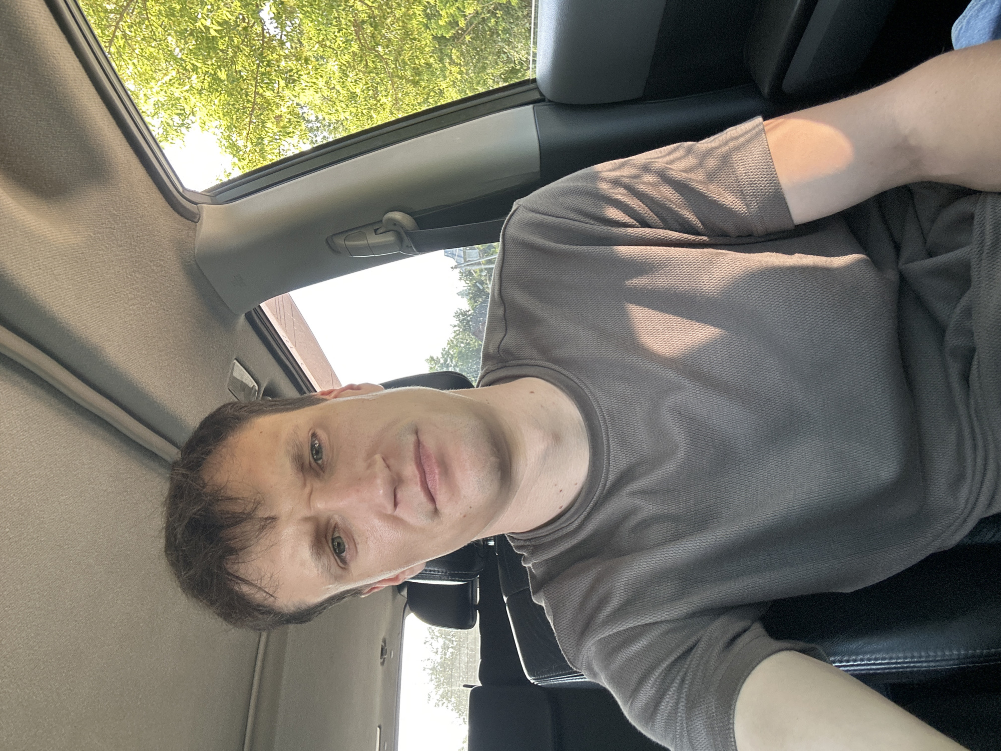 A man sits in the driver's seat of a car, looking at the camera. He has short, brown hair and is wearing a brown t-shirt. Sunlight streams in from the right side of the car, illuminating the man's arm and casting shadows on his shirt. The interior of the car is visible, including the headrests of the seats behind him and the car's ceiling. Outside the car, green trees and a bright sky are visible through the window. The location is Makiv, Ukraine.