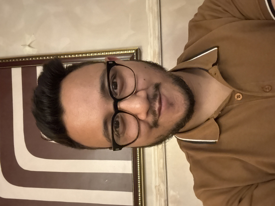 A person wearing glasses and a brown shirt stands next to a framed, geometric art piece. The shirt has a contrasting white trim on the collar. The art piece features alternating brown and white curved stripes. The person's face is visible, and they have a dark beard and short, dark hair. The background includes a plain white wall with a small, torn area. The image is lit softly, creating subtle shadows.