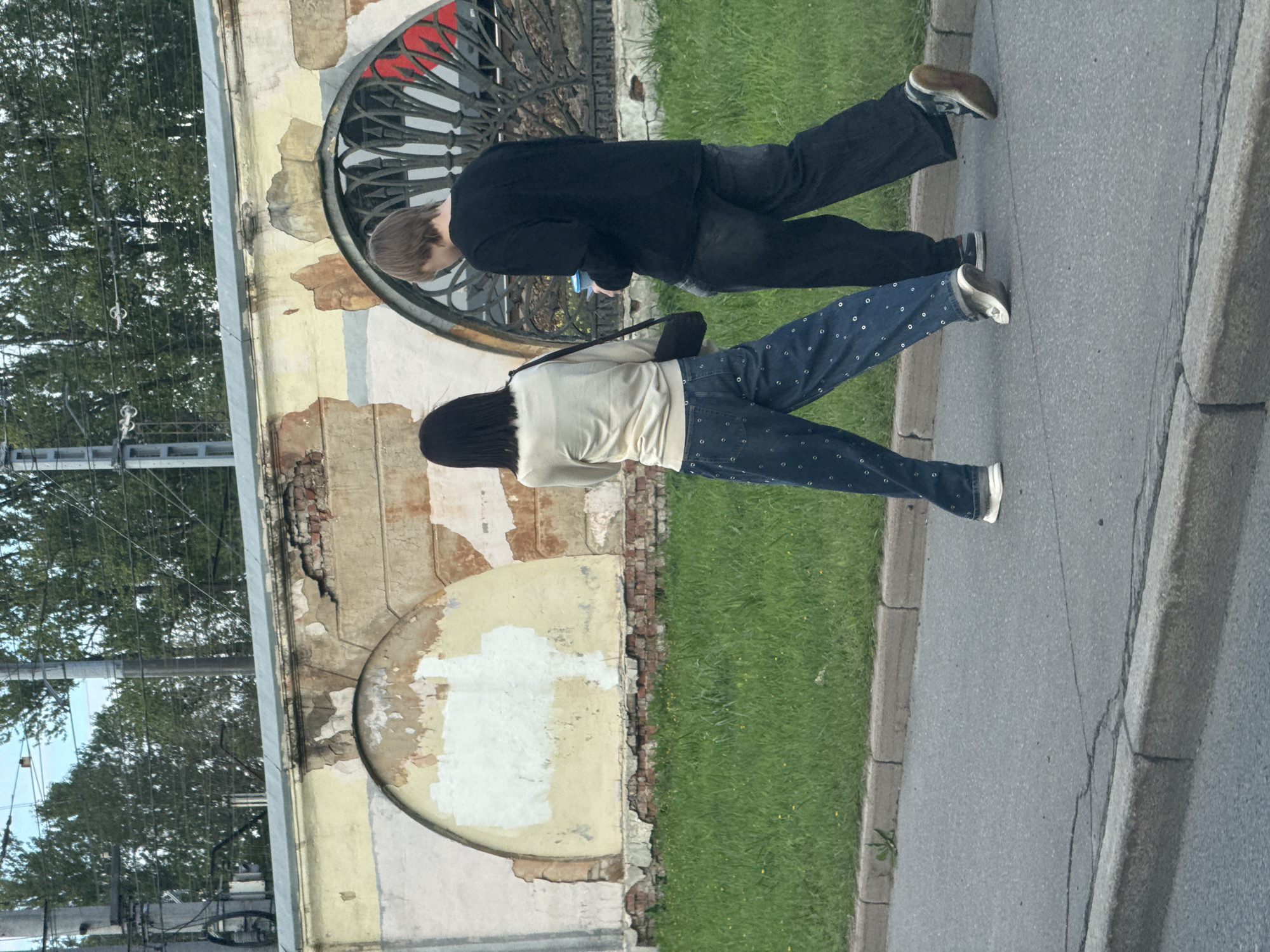 Two individuals, a woman with dark hair and a man with light brown hair, walk along a sidewalk adjacent to a grassy area. The woman wears a light-colored, long-sleeved top and wide-leg jeans with a patterned design. She carries a dark crossbody bag. The man wears a black long-sleeved shirt and dark pants. He holds a blue cup in his right hand. Behind them is an old, weathered wall with peeling paint and brickwork, featuring arched recesses and an ornate metal grate. The environment appears to be an urban setting in Saint Petersburg, Russia, suggested by the style of the architecture and the presence of overhead power lines and poles, typical of a city's infrastructure. The ground is paved with asphalt and bordered by a concrete curb. The overall scene depicts a casual street view with architectural decay and everyday activity.