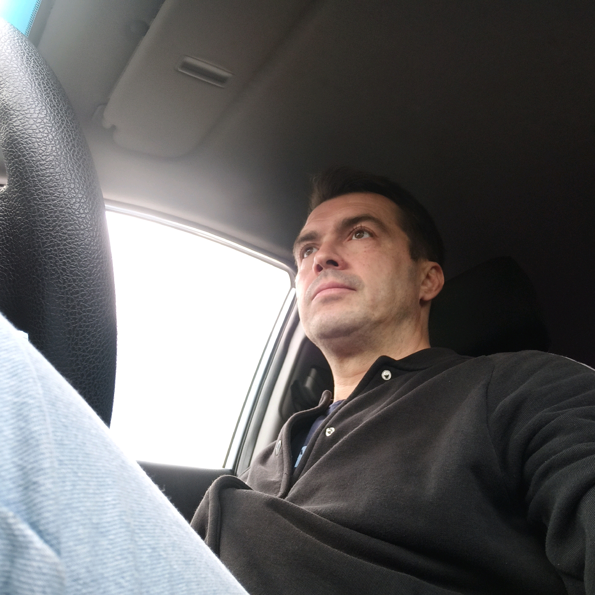 The image shows a man sitting in the driver's seat of a car, looking upwards and to his right. He is wearing a dark long-sleeved shirt with a light blue collar peeking out from underneath, and light blue jeans. The interior of the car is visible, including the steering wheel on the left side of the frame and the interior roof lining. The sun visor is down, and the passenger side window is visible, showing a bright, overcast sky. The image appears to be a selfie taken by the man. The scene is generic and does not provide any specific details that would indicate a location within Nizhny Novgorod, Russia.